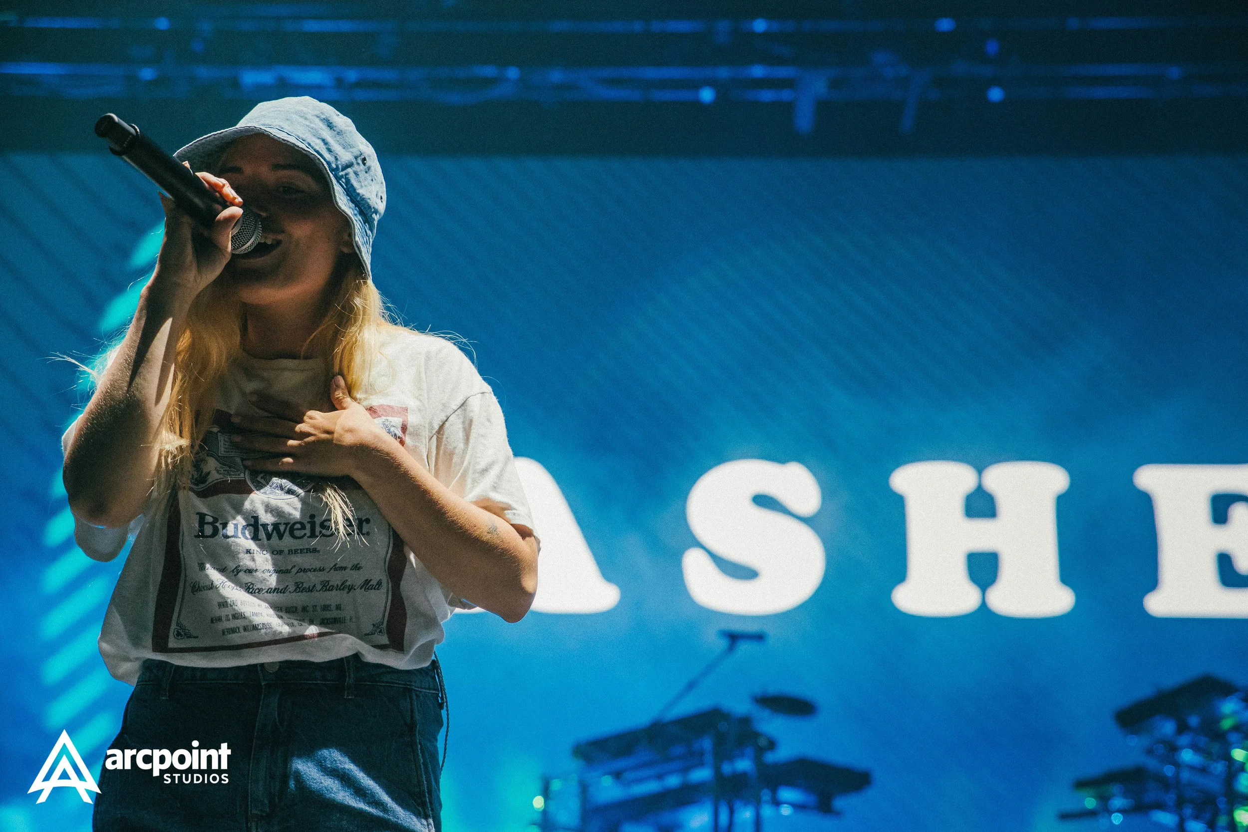 A woman with long blonde hair singing into a microphone on stage, wearing a light denim bucket hat and a white T-shirt with a Budweiser logo, with her hand on her chest, illuminated by stage lighting, with large white text in the background and the A