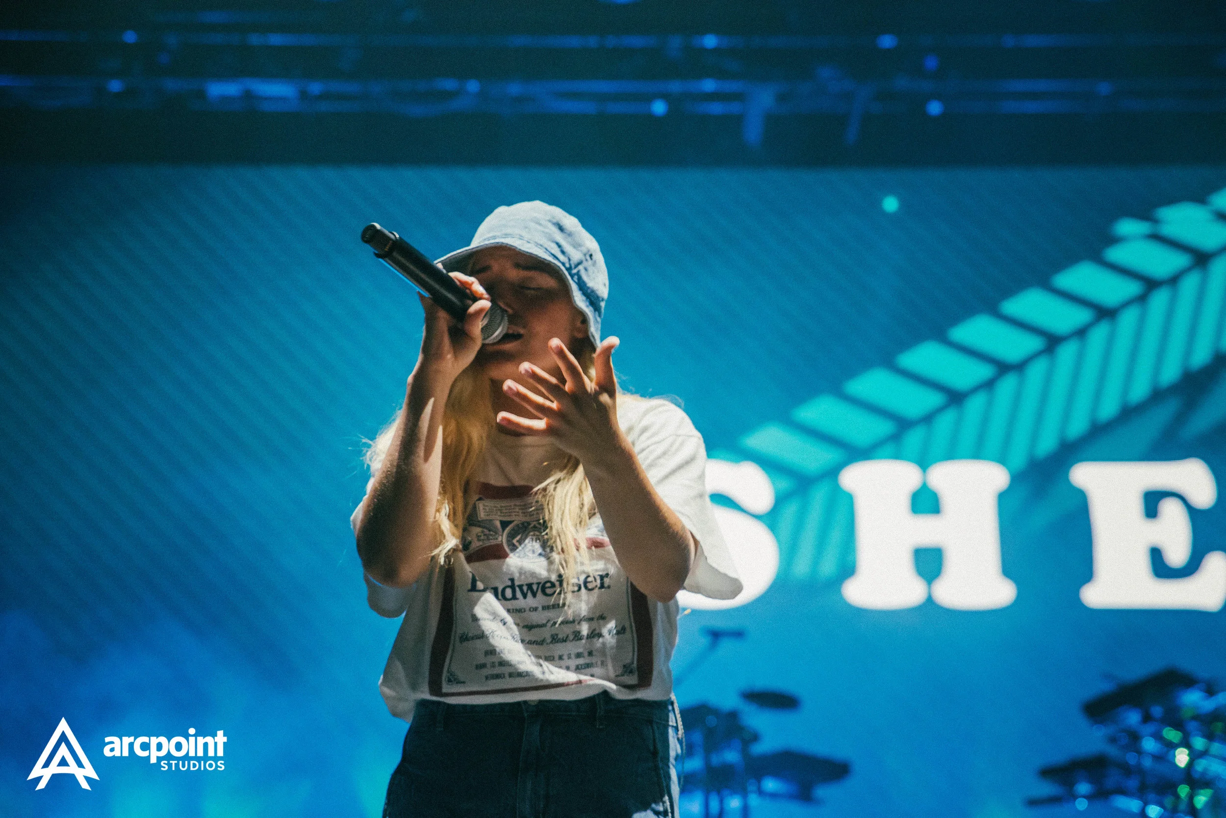 A female singer wearing a bucket hat performs on stage with a microphone, surrounded by stage lights and a large screen in the background.