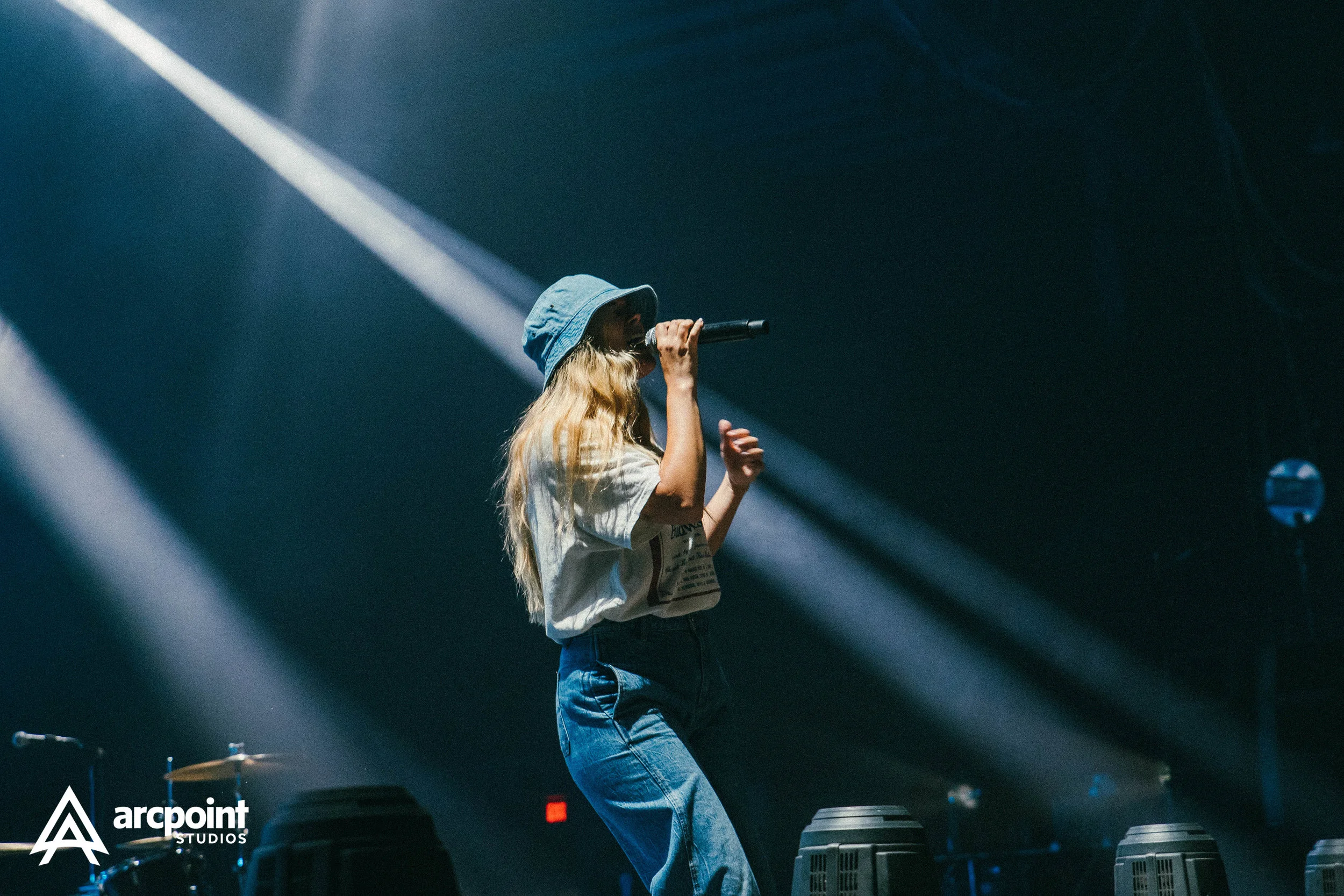 A female singer with long blonde hair, wearing a blue bucket hat and casual clothing, performs on stage with a microphone, with stage lighting and musical equipment in the background.
