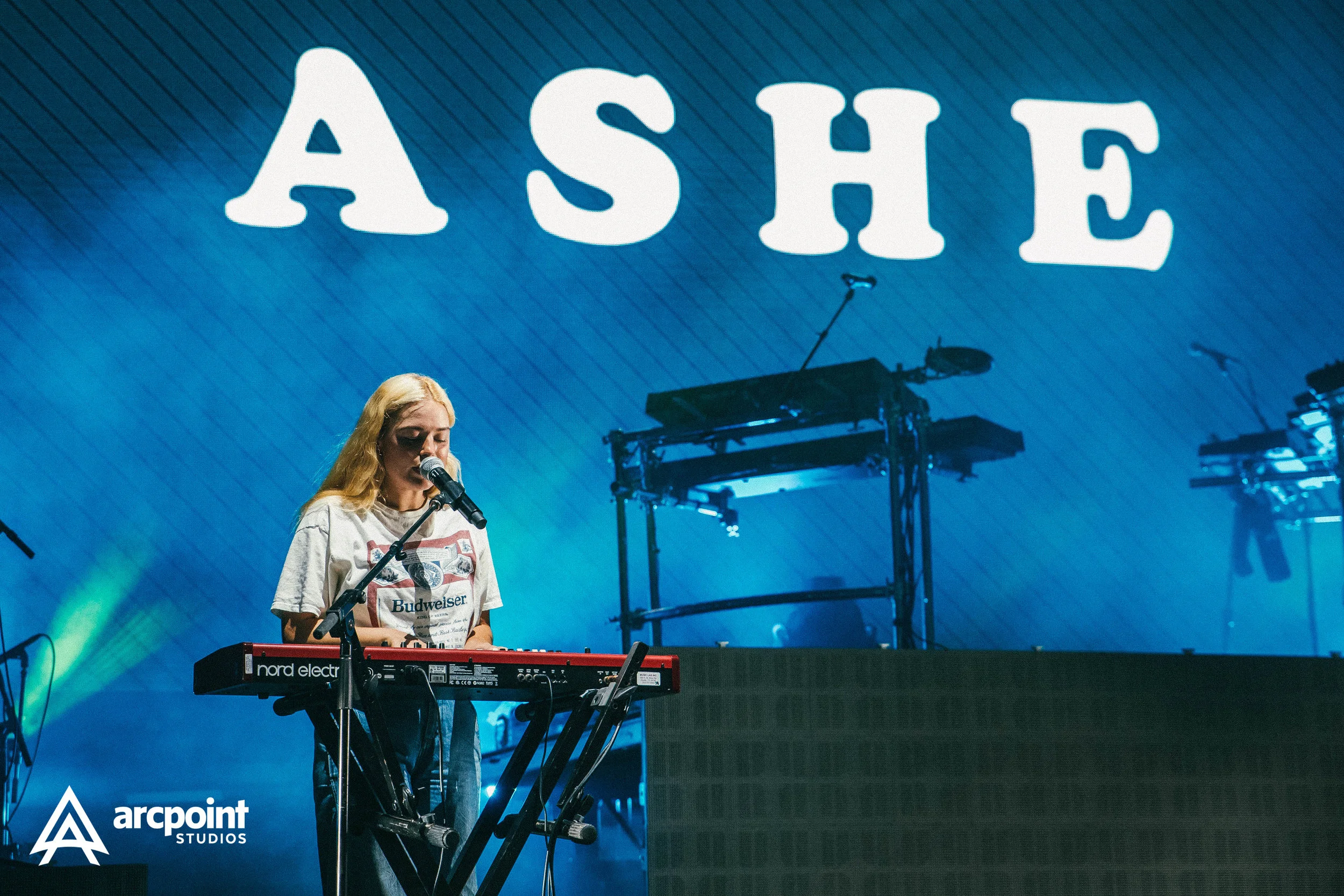A young female musician performing on stage, singing into a microphone while playing a red Nord electric keyboard, with a large illuminated sign spelling 'ASHE' in the background and blue stage lighting.
