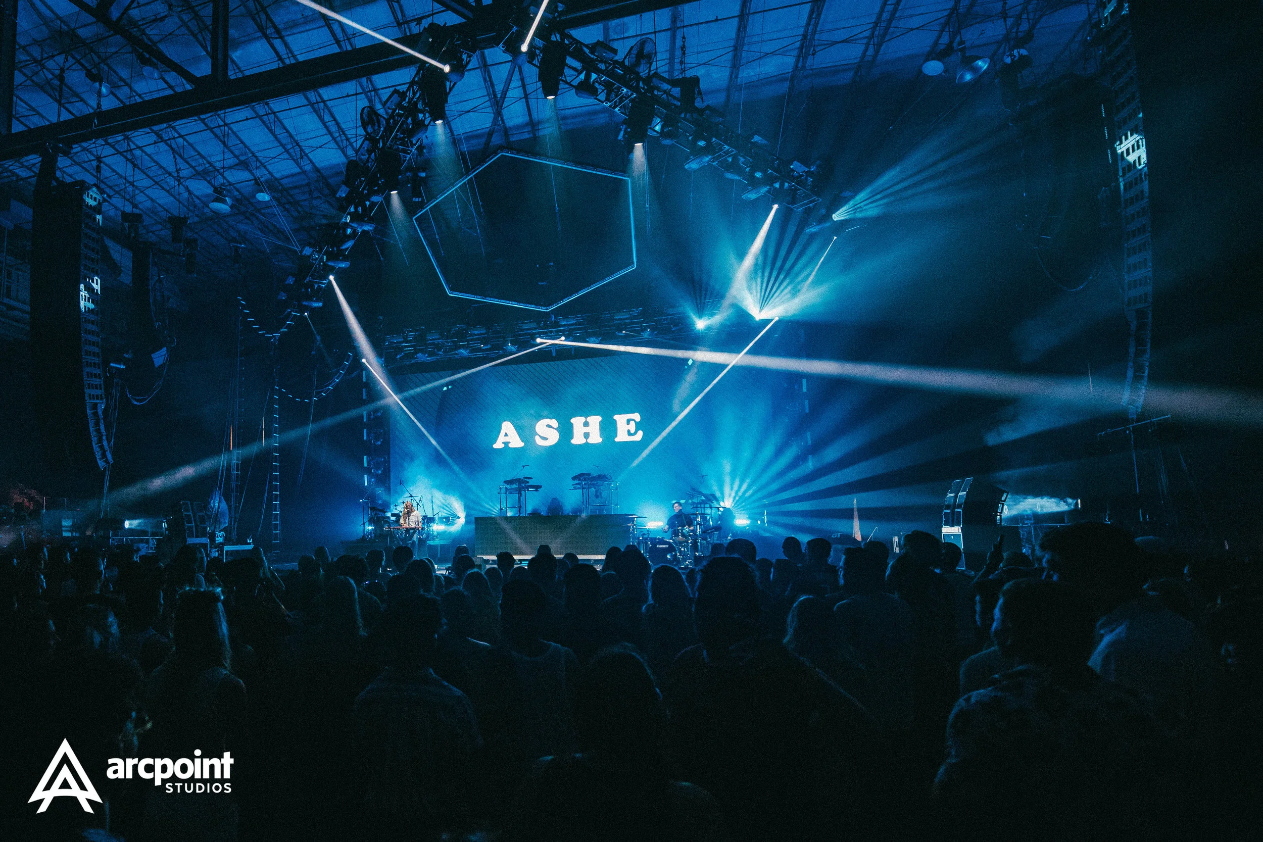 A concert stage with blue lighting displaying the word 'ASHE' on a large screen. There are musicians and electronic equipment on stage, with beams of light directed toward the audience. The foreground shows a crowd of people watching the performance 