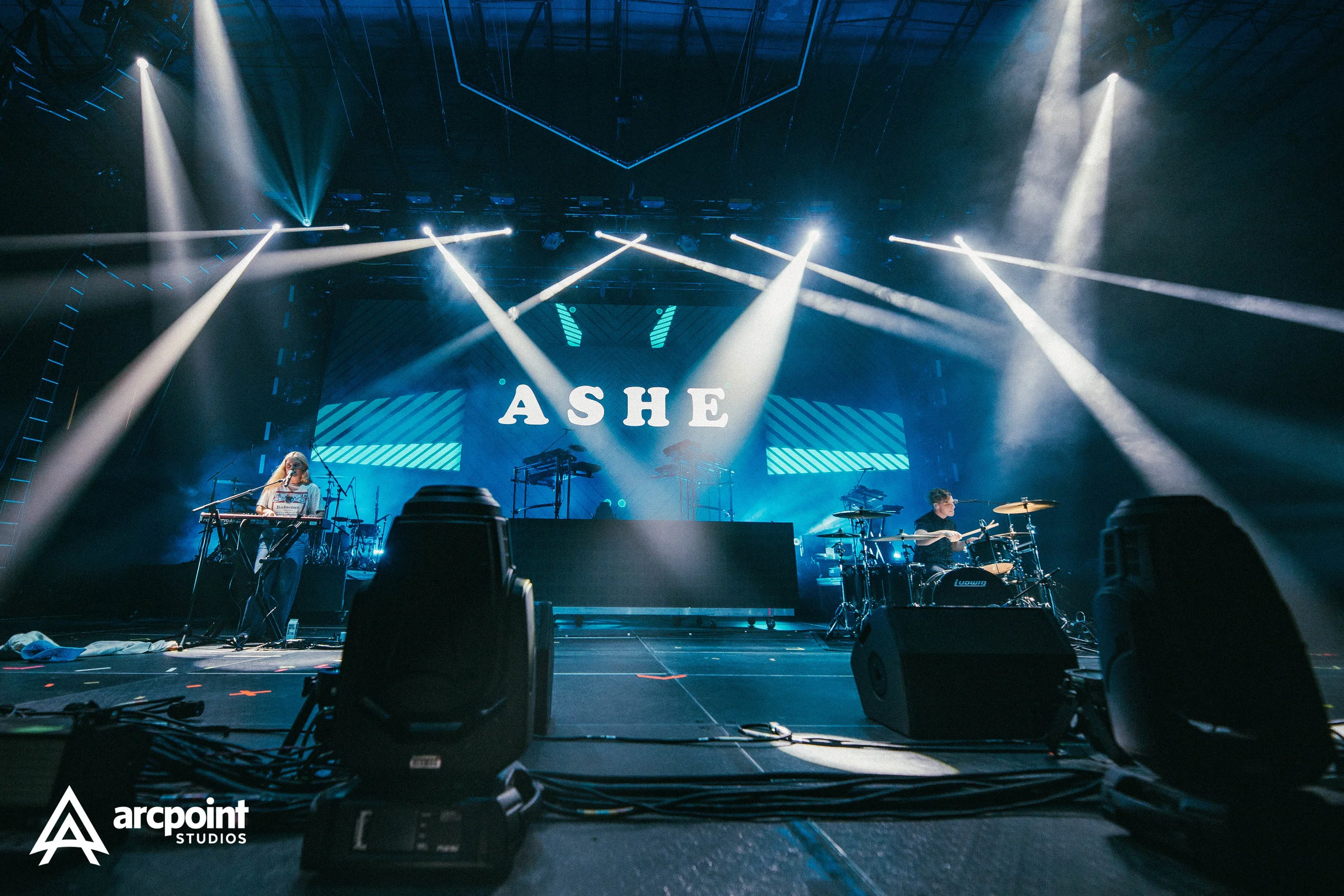 Stage with a large screen displaying the word 'ASHE,' illuminated by multiple stage lights and spotlights, with two musicians playing keyboards and drums.