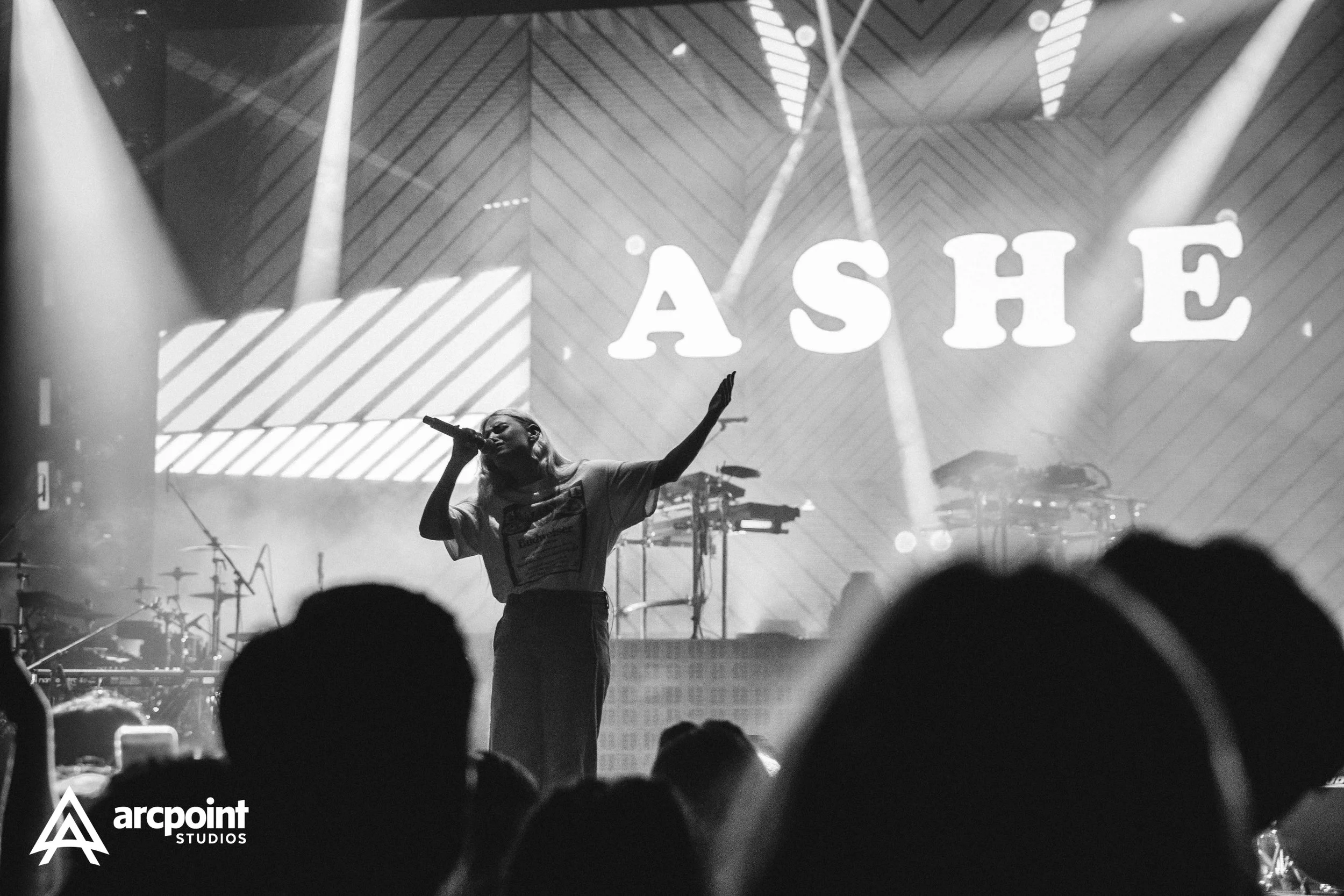 A black and white photo of a woman singing into a microphone on stage with the word "ASHE" displayed in large letters behind her and the logo "arcpoint STUDIOS" in the bottom left corner.