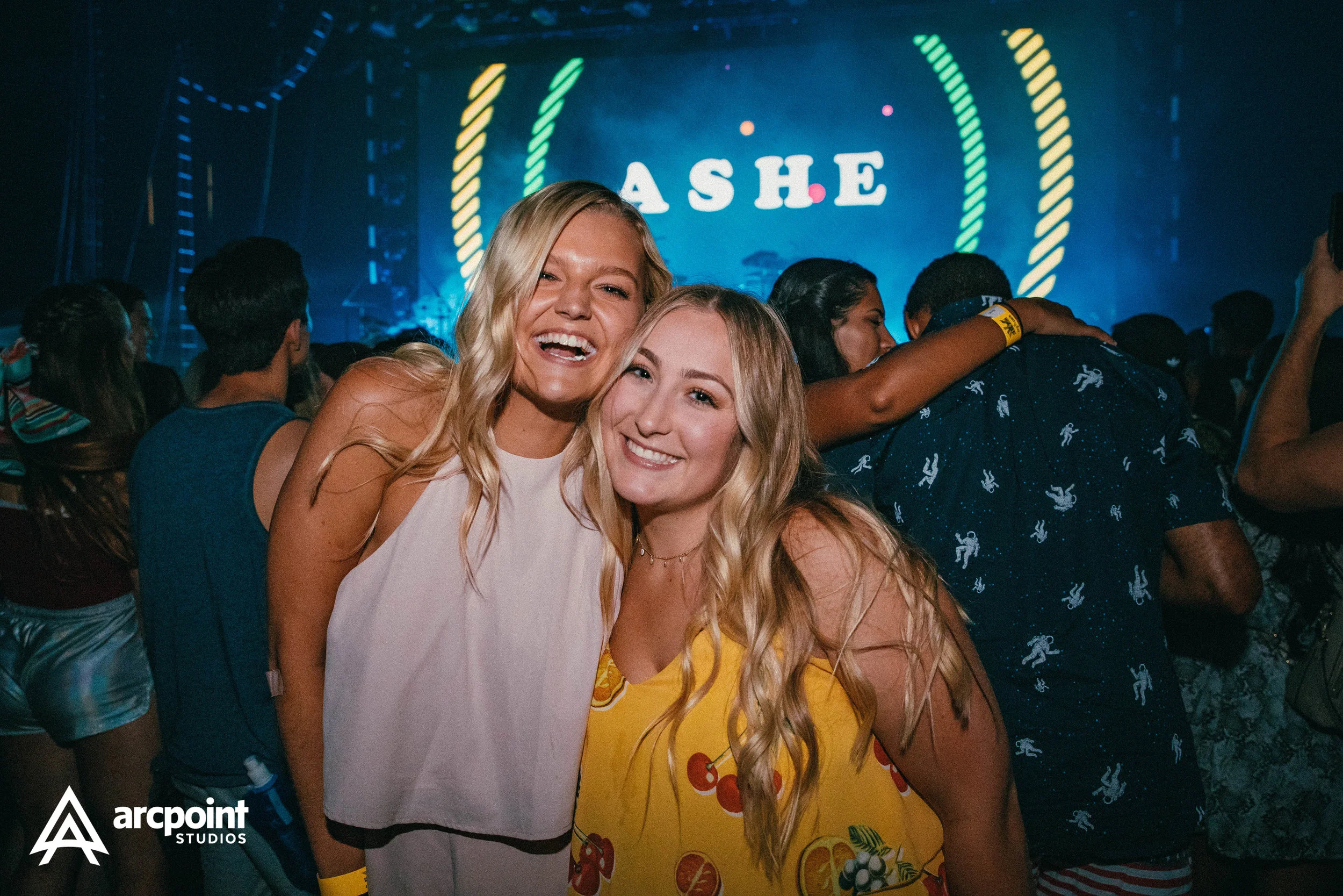 Two women smiling and posing for the camera at a concert or event, with a stage in the background displaying the word 'ASHE'.