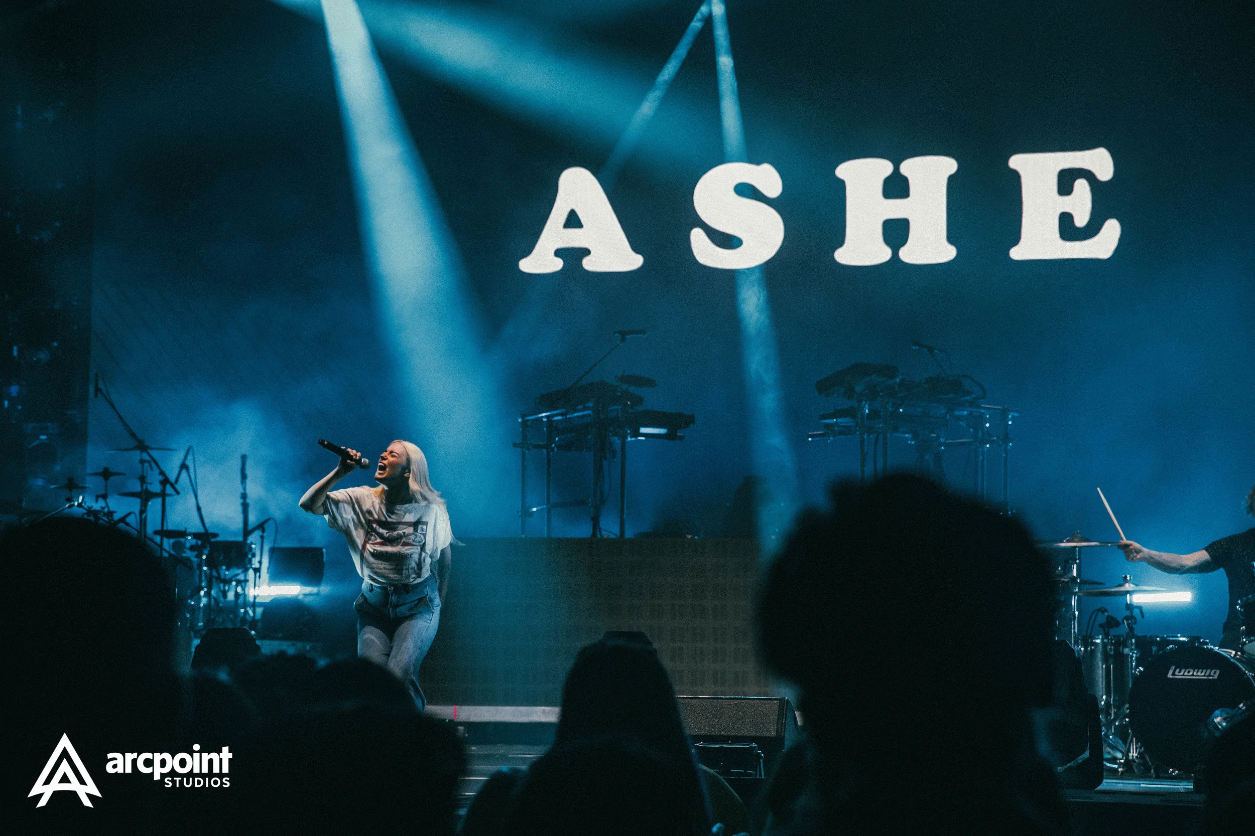 A woman singing on stage with a microphone, with the word "ASHE" projected behind her, and a band playing in the background. Audience silhouettes are visible in the foreground.