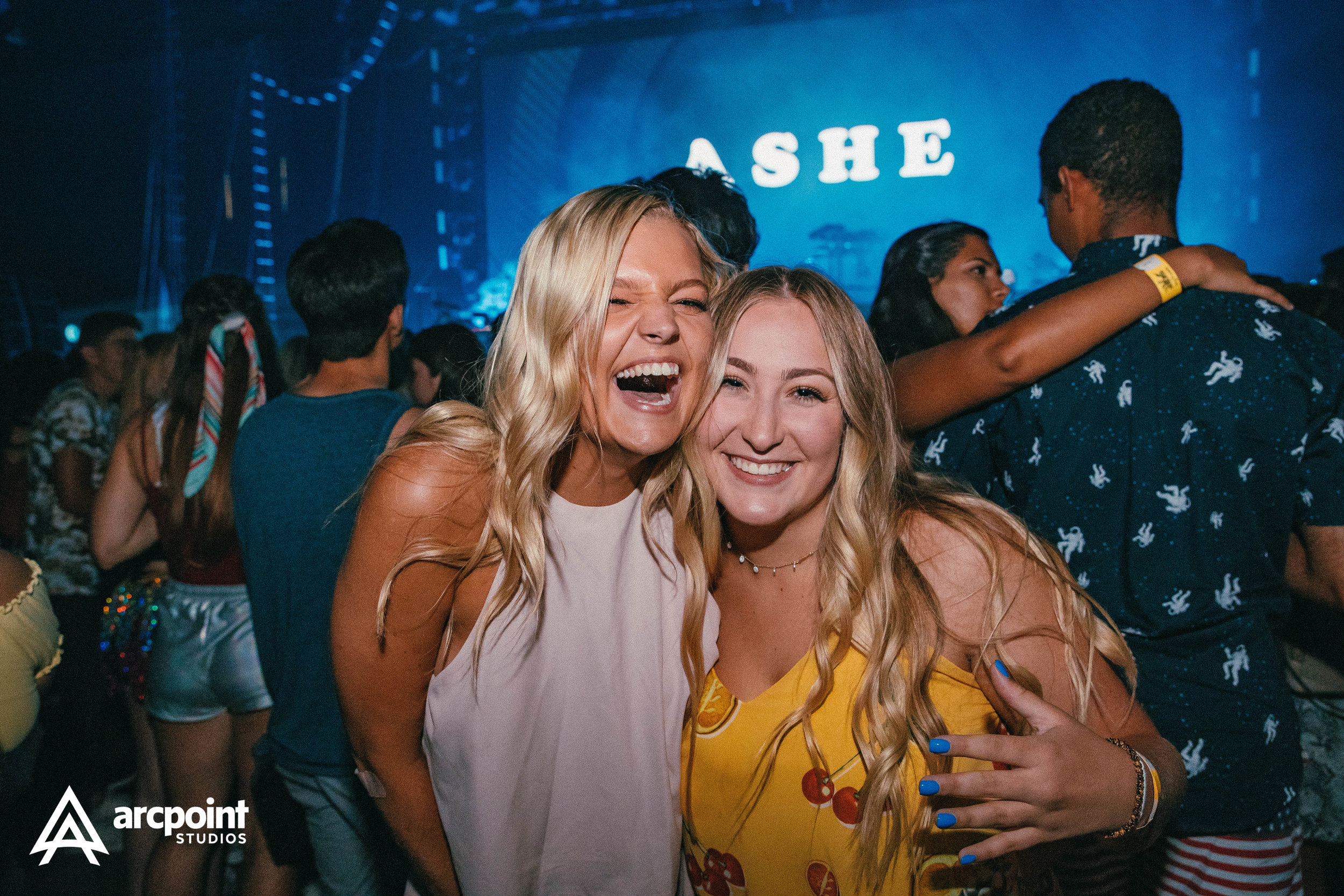 Two young women smiling and hugging at a crowded event with a stage in the background displaying the word 'ASHE'.