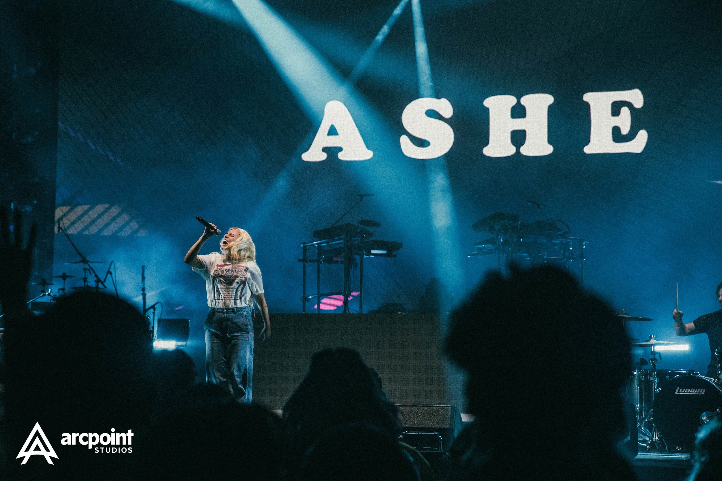 A female singer performing on stage with a microphone, with the word "ASHE" projected on a large screen behind her, and a drummer visible to the right. The stage is lit with blue lighting.