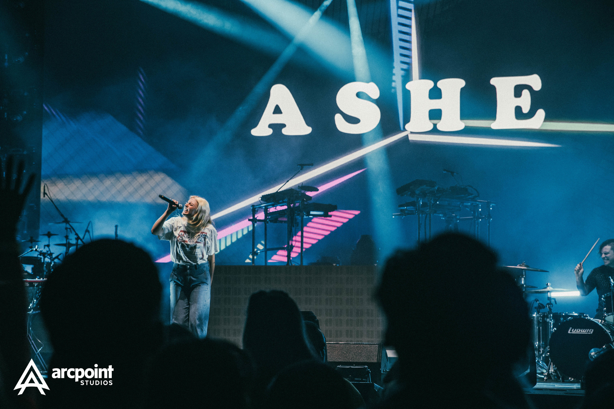 A live concert scene with a female singer holding a microphone on stage, accompanied by a drummer. The backdrop features a large illuminated sign spelling 'ASHE' with stage lights and electronic equipment around.