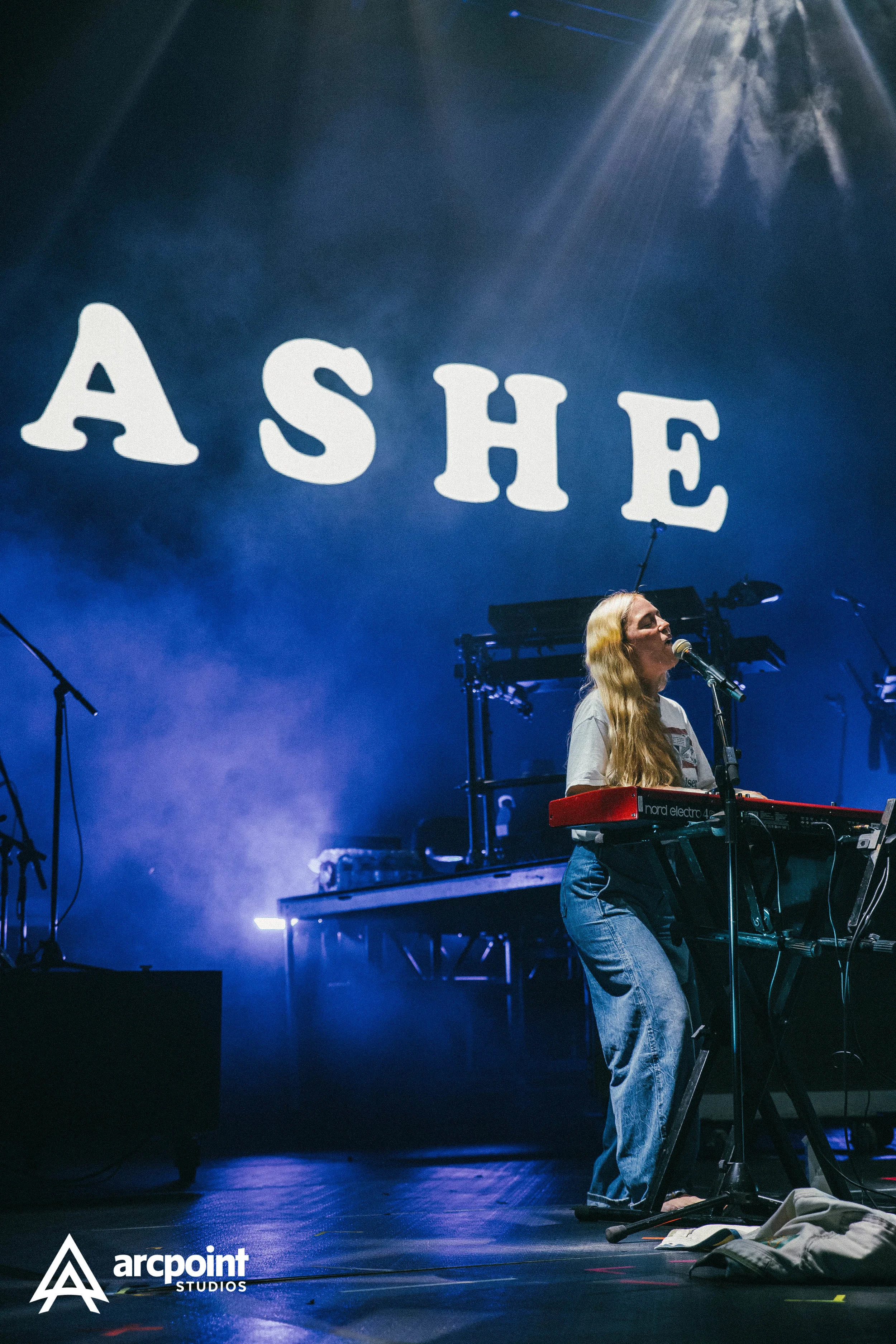 A woman playing a keyboard on stage during a concert, with the word 'ASHE' projected in large letters on the backdrop.