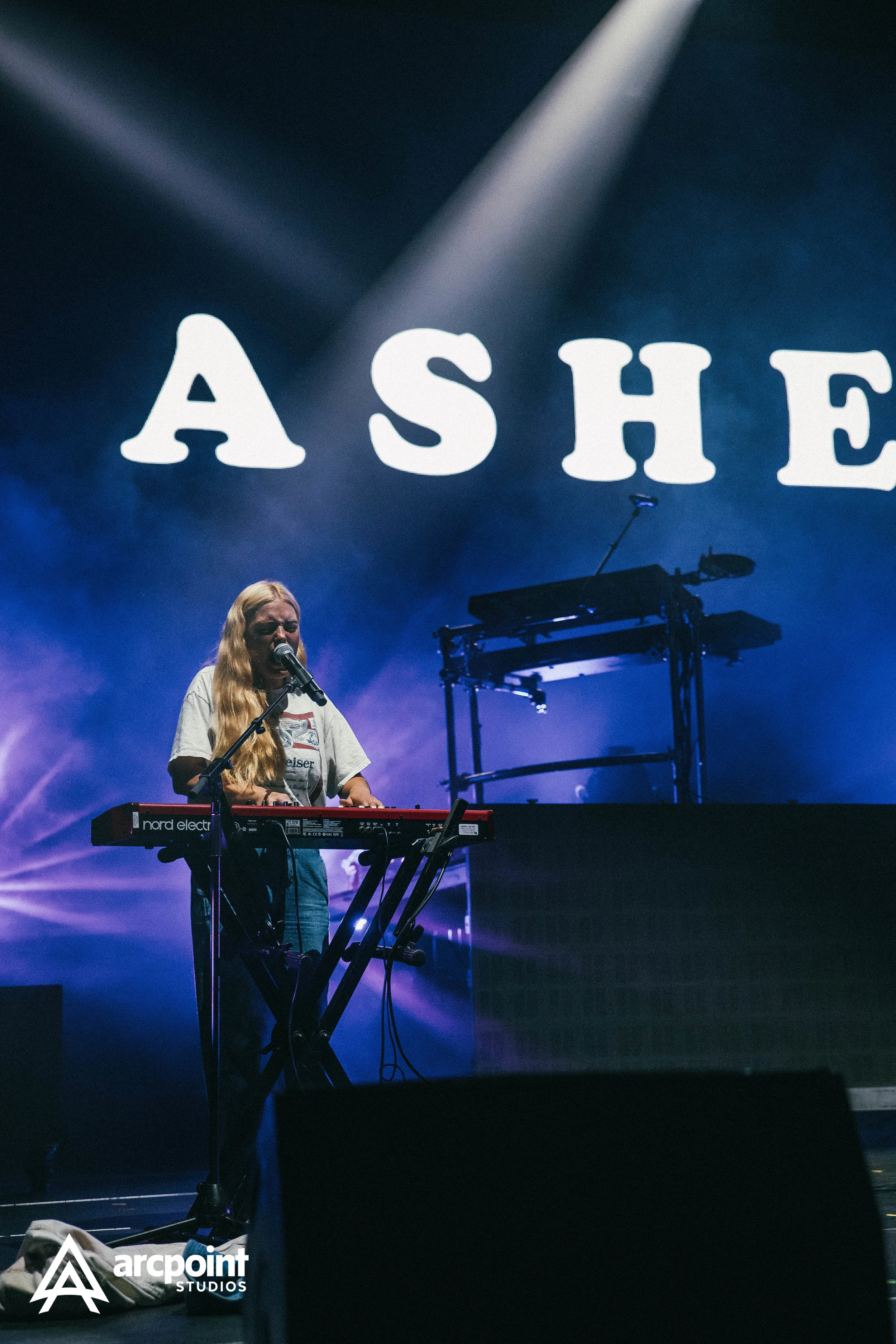 A woman with long blonde hair singing into a microphone while playing a keyboard on stage during a concert, with a large illuminated screen behind her displaying the word 'ASHE' and stage lights creating a purple and blue atmosphere.