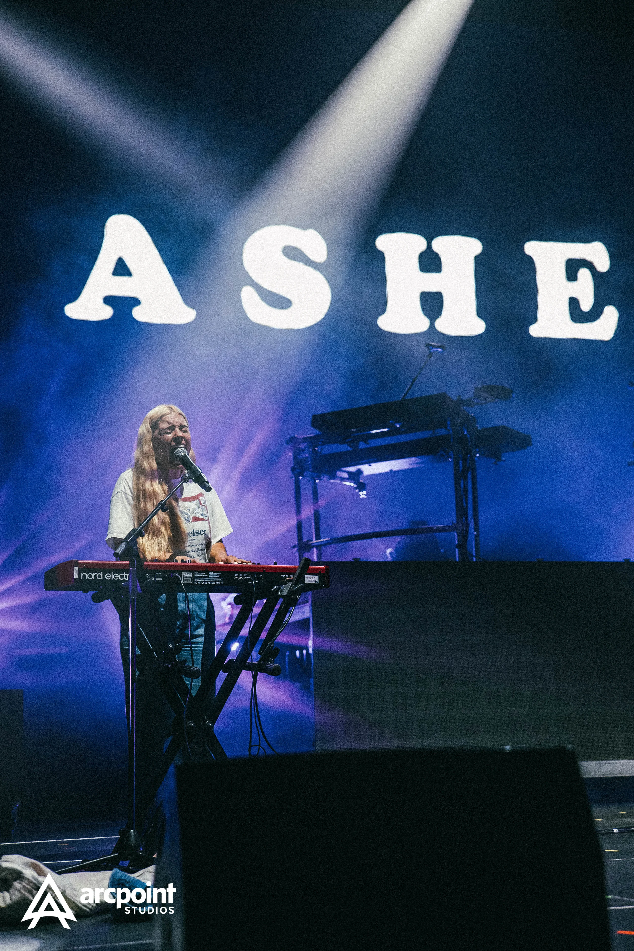 A woman performs on stage at a concert, singing into a microphone and playing a red keyboard. Behind her, large illuminated letters spell 'ASHE'. Stage lights cast purple and blue hues.
