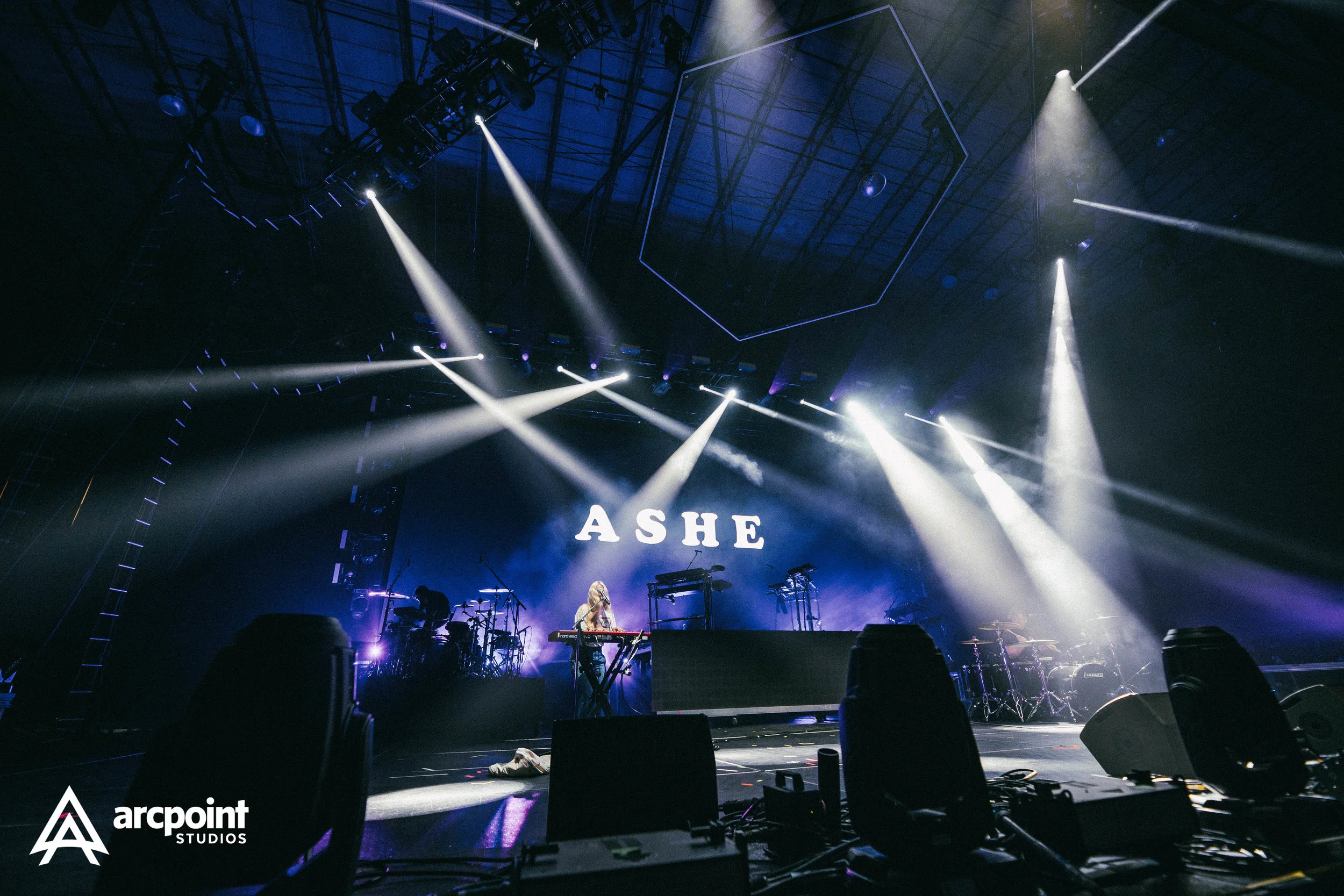 A concert stage with bright white stage lights, a musician playing keyboard, and the word "ASHE" displayed on a large screen behind the performer. Several musical instruments and equipment are visible, with the logo "arcpoint STUDIOS" in the bottom l