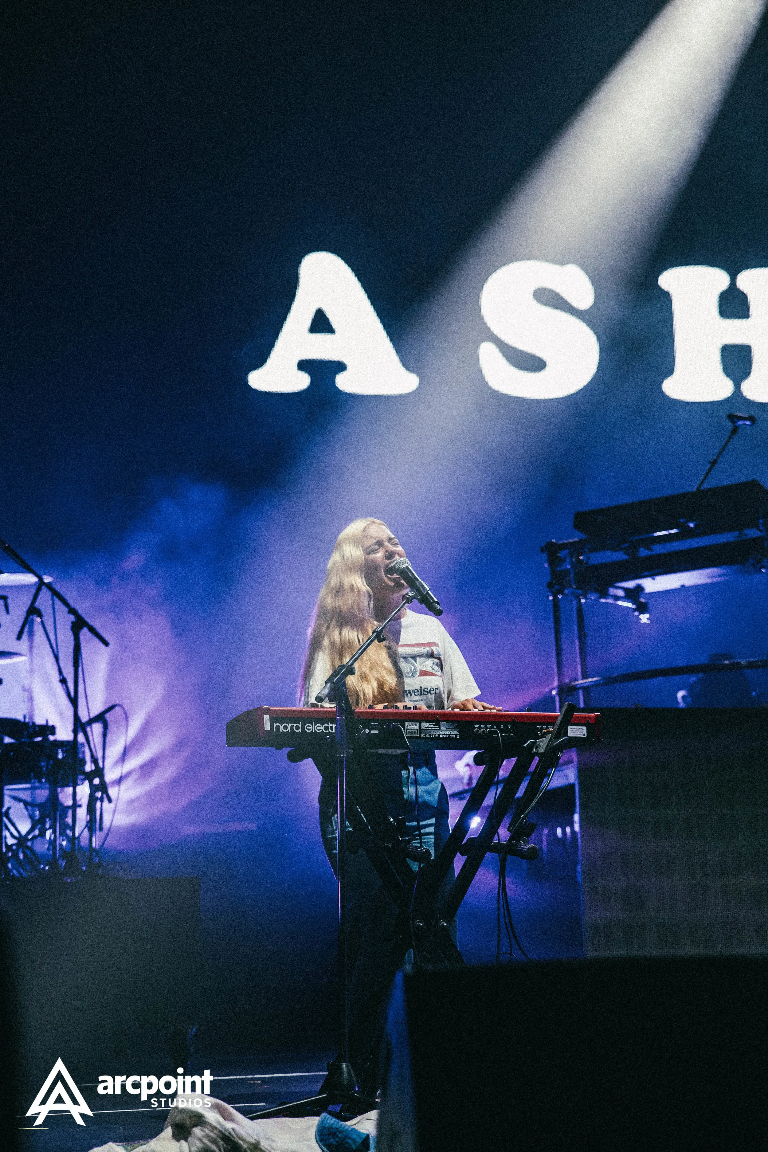 A female singer performs on stage with a keyboard, microphone, and background screen displaying large, white letters.