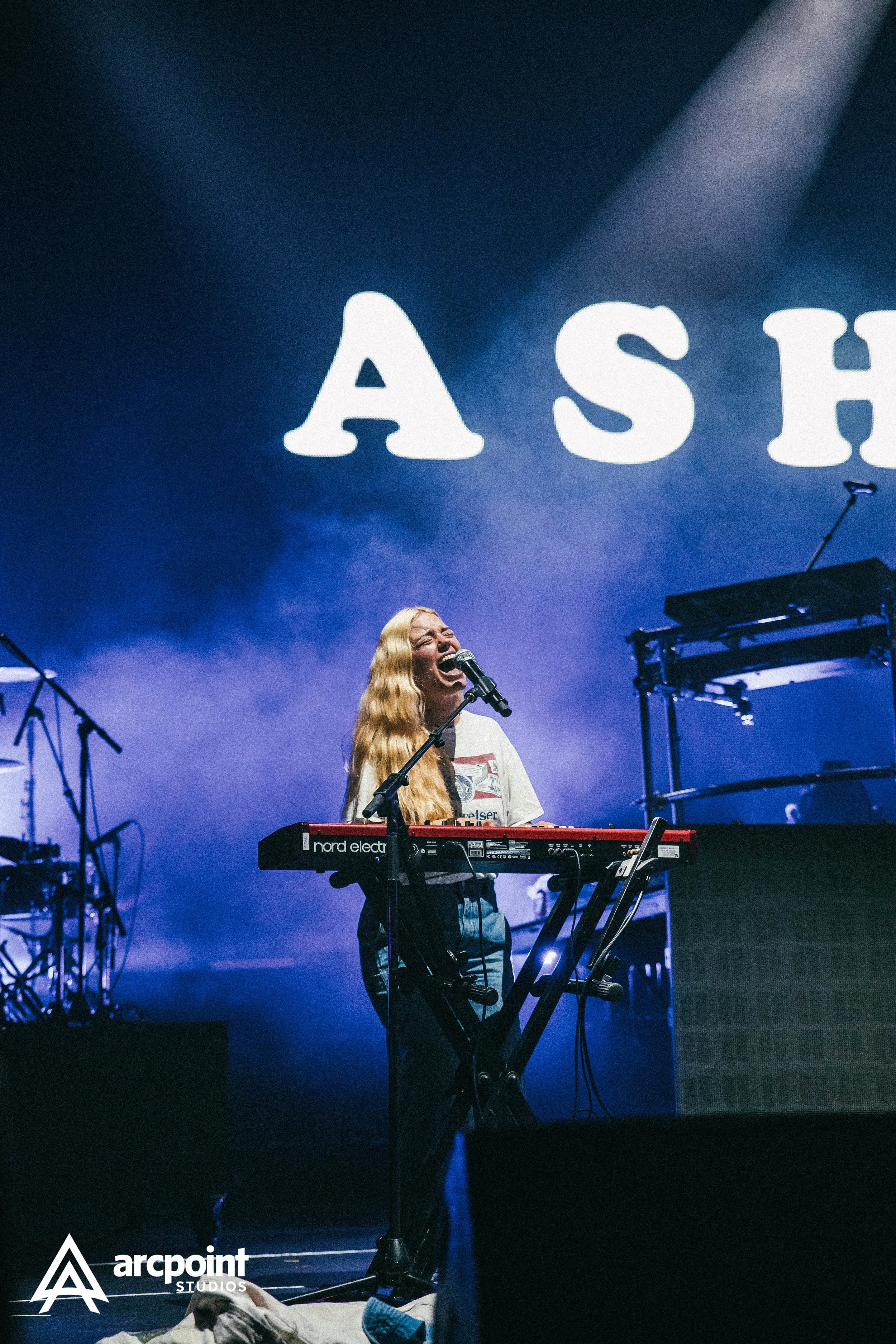 A woman with long blonde hair singing into a microphone while playing a keyboard on stage, with large white letters spelling 'ASH' projected behind her.