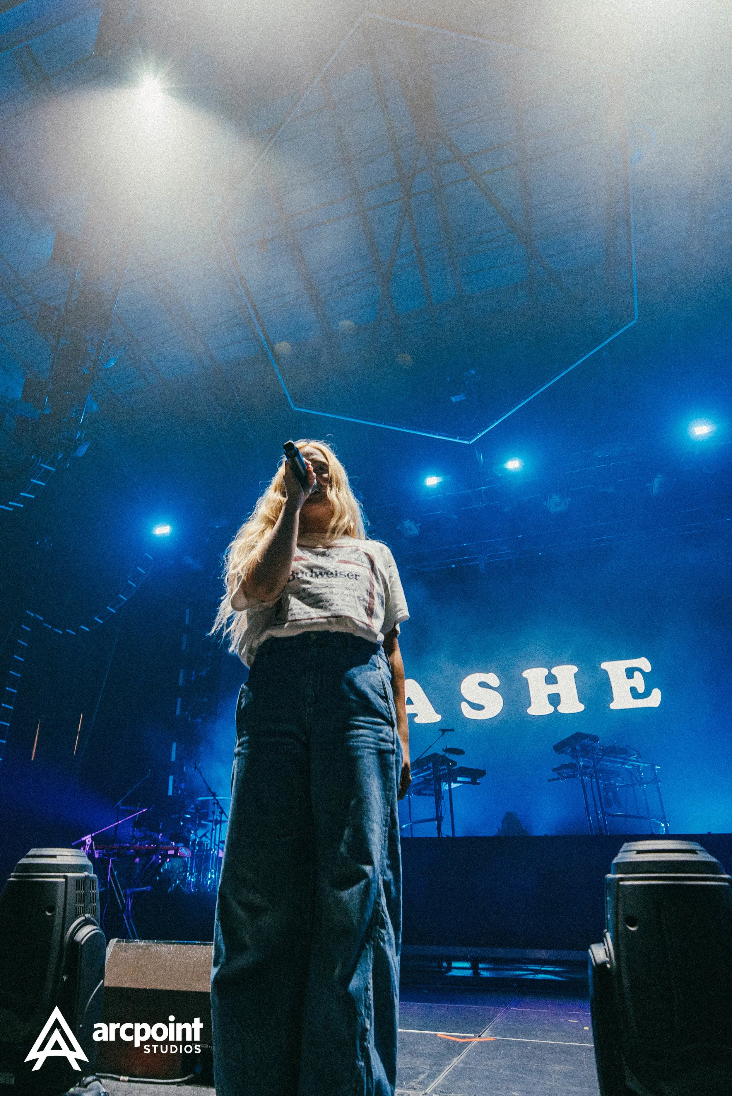 A young woman with long blonde hair singing into a microphone on stage with blue lights and a large screen displaying 'ASHE' behind her. She is wearing a graphic T-shirt and high-waisted jeans, and there are electronic music equipment and speakers on