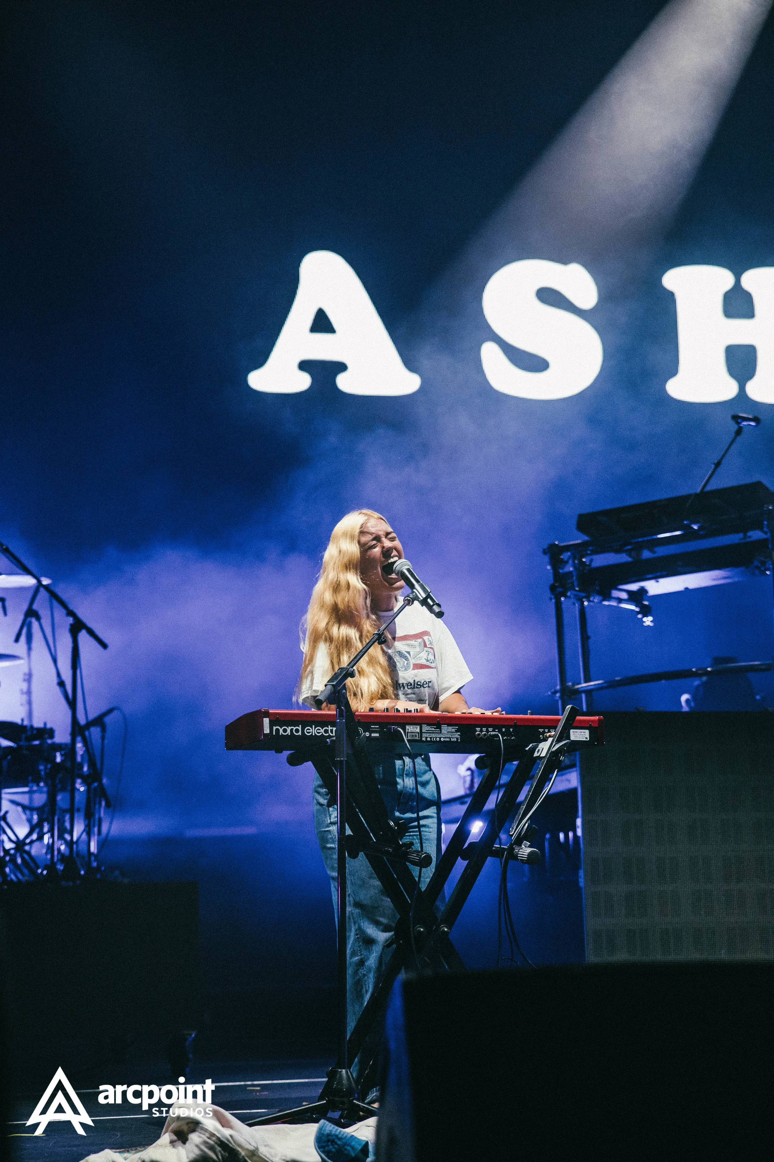 A woman with long blonde hair singing passionately into a microphone while playing a red Nord Electro keyboard on stage with blue and purple lighting and large white letters displaying 'ASH' in the background.