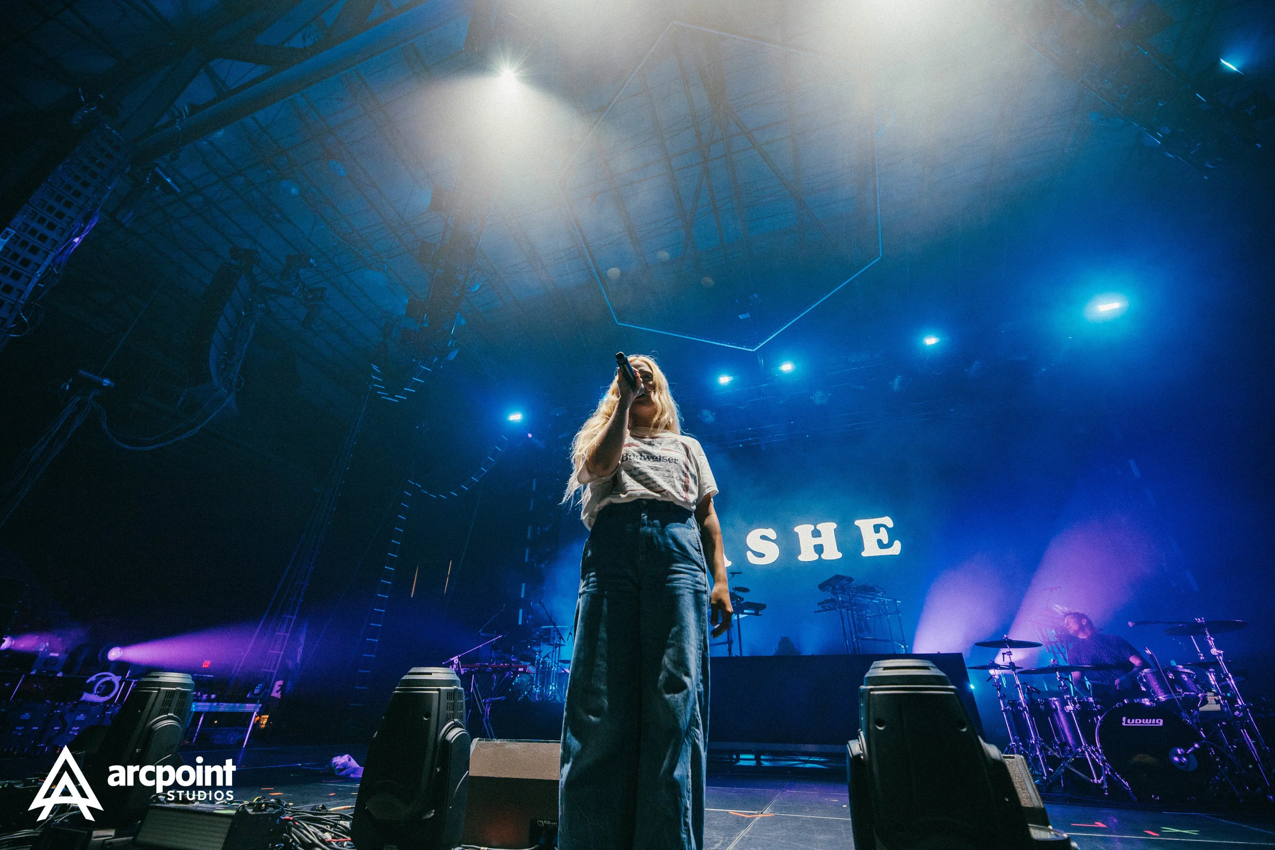 A female singer performs on stage with a microphone, wearing a t-shirt and wide-leg jeans, with blue stage lights and a drum set in the background, and the word 'SHE' projected on the backdrop.