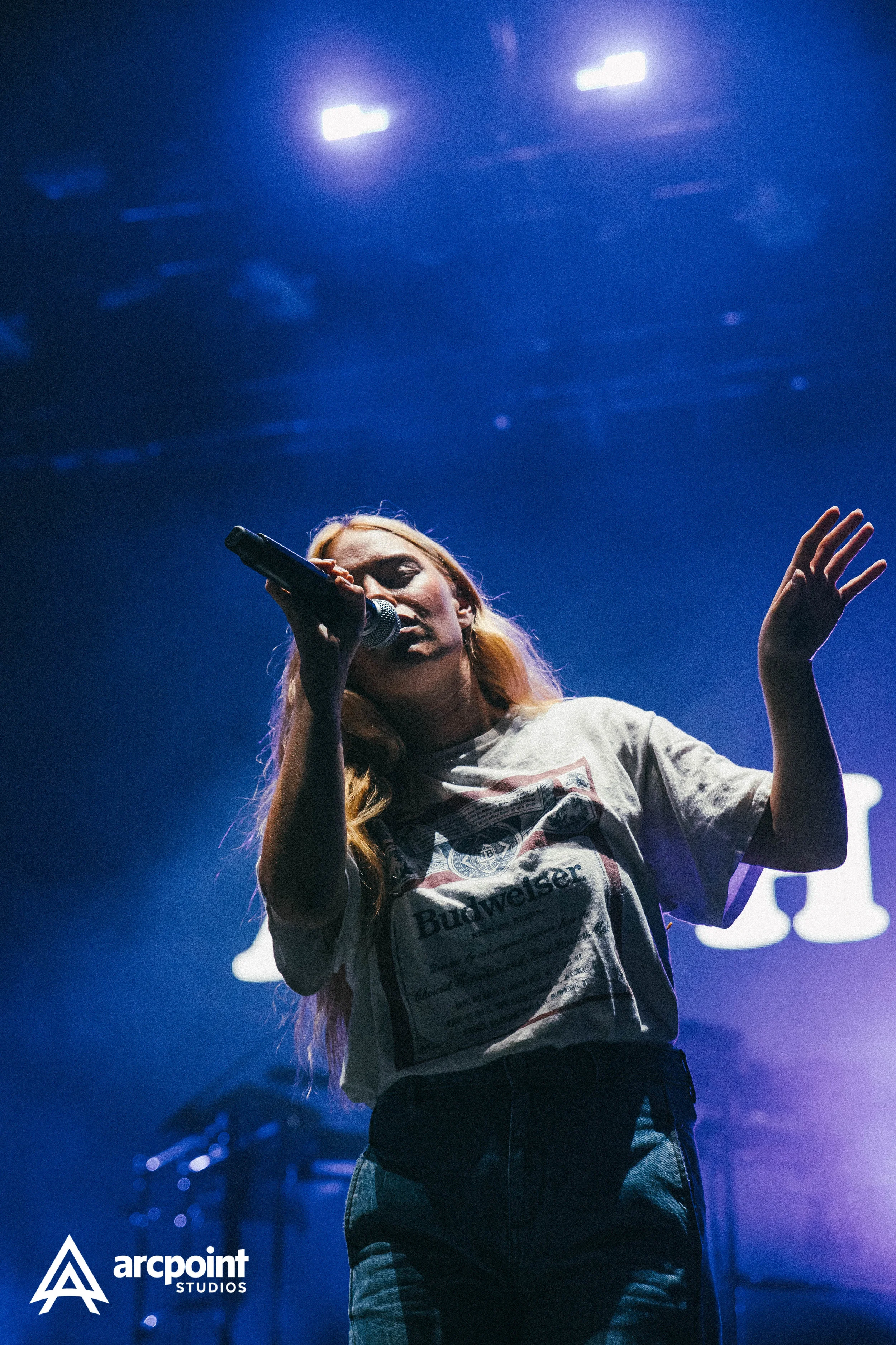 A woman singing into a microphone on a stage with blue lighting, wearing a graphic t-shirt and jeans.