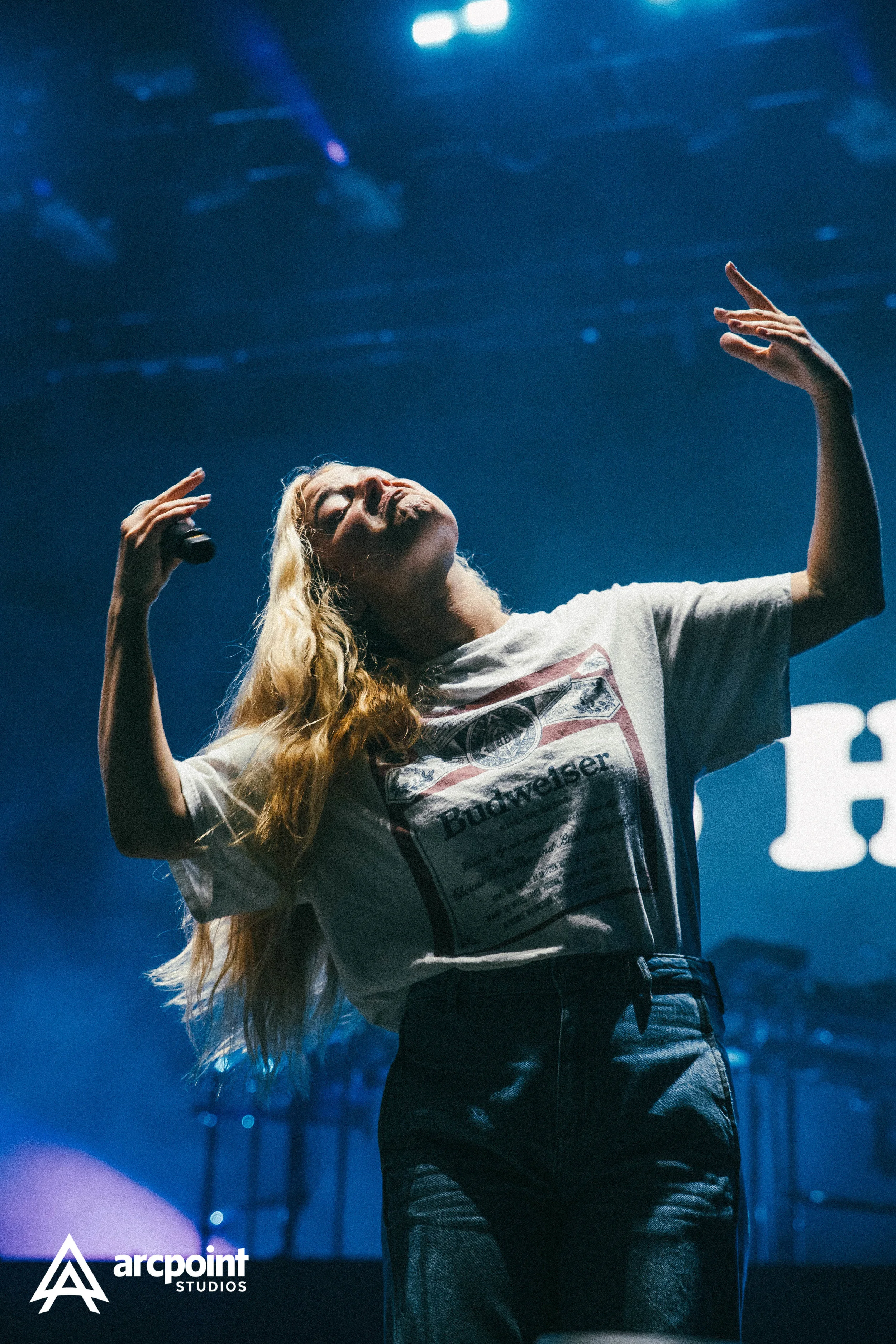 A woman with long blonde hair singing or performing on stage with her eyes closed and hands raised in the air, wearing a white T-shirt with a Budweiser logo and dark high-waisted jeans, under blue stage lighting.