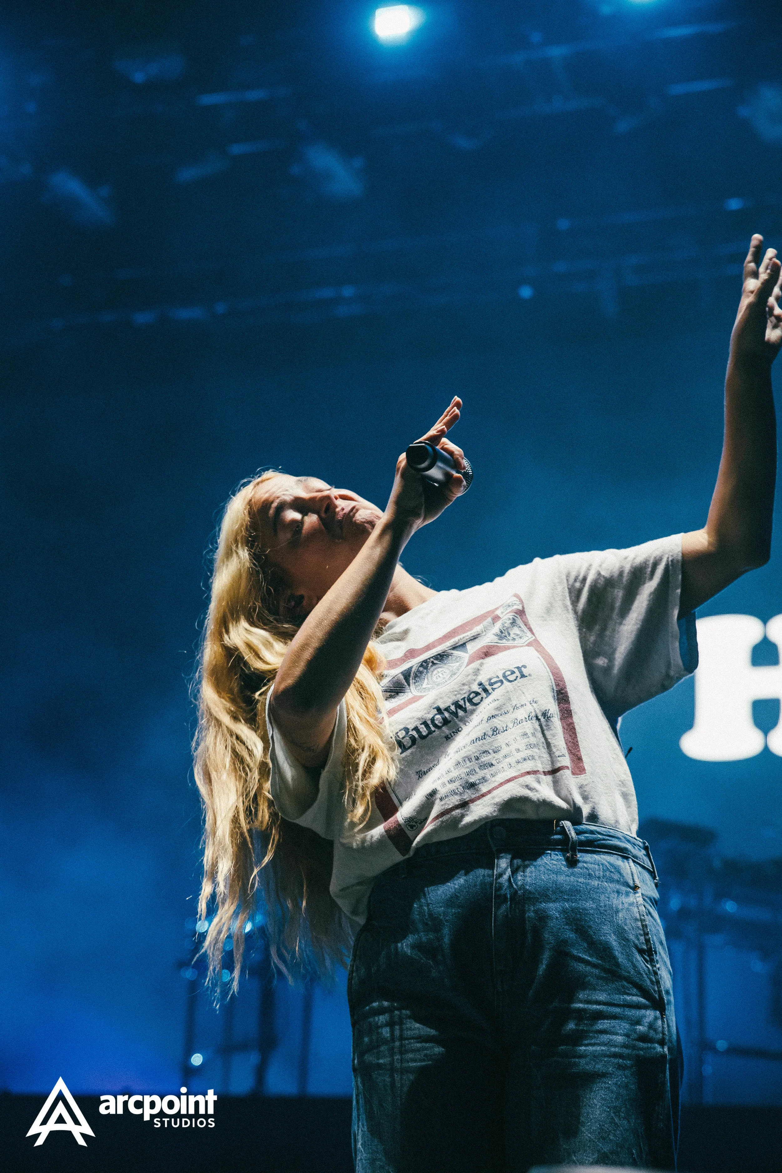 A female singer with long blonde hair performing on stage with a microphone in hand and a large screen with a logo in the background. She is wearing a vintage Budweiser t-shirt and jeans.