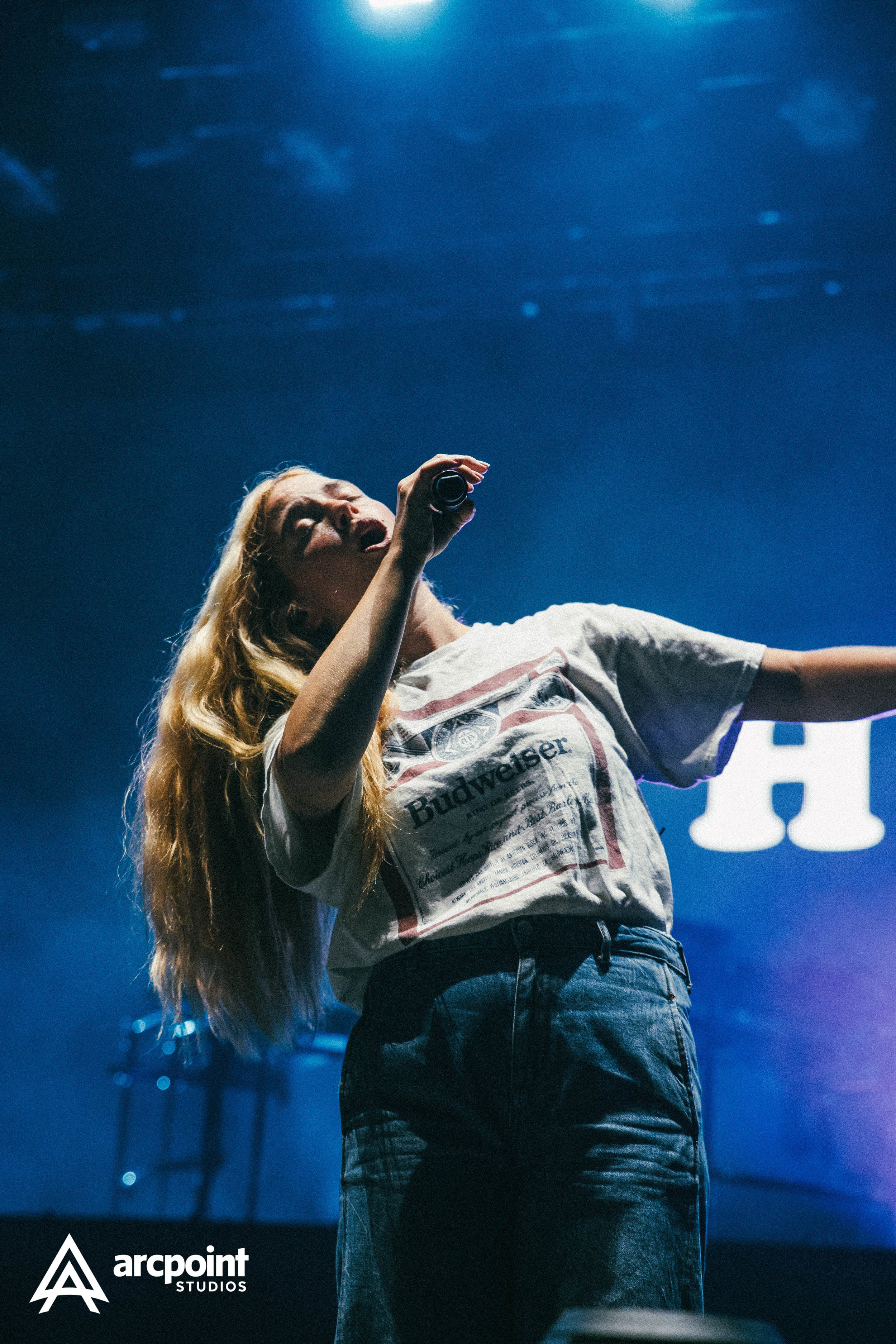 A woman with long blond hair singing into a microphone on stage with blue lighting in the background, wearing a gray T-shirt.