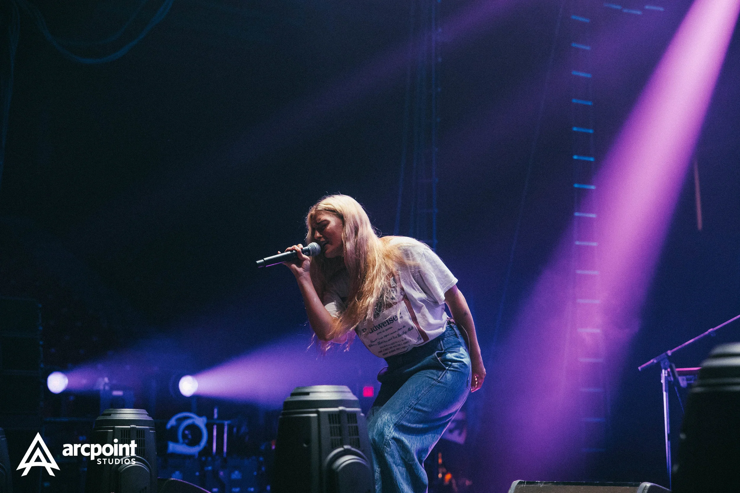 Female singer performing on stage with purple and blue lighting, holding a microphone, wearing a white t-shirt and blue jeans.
