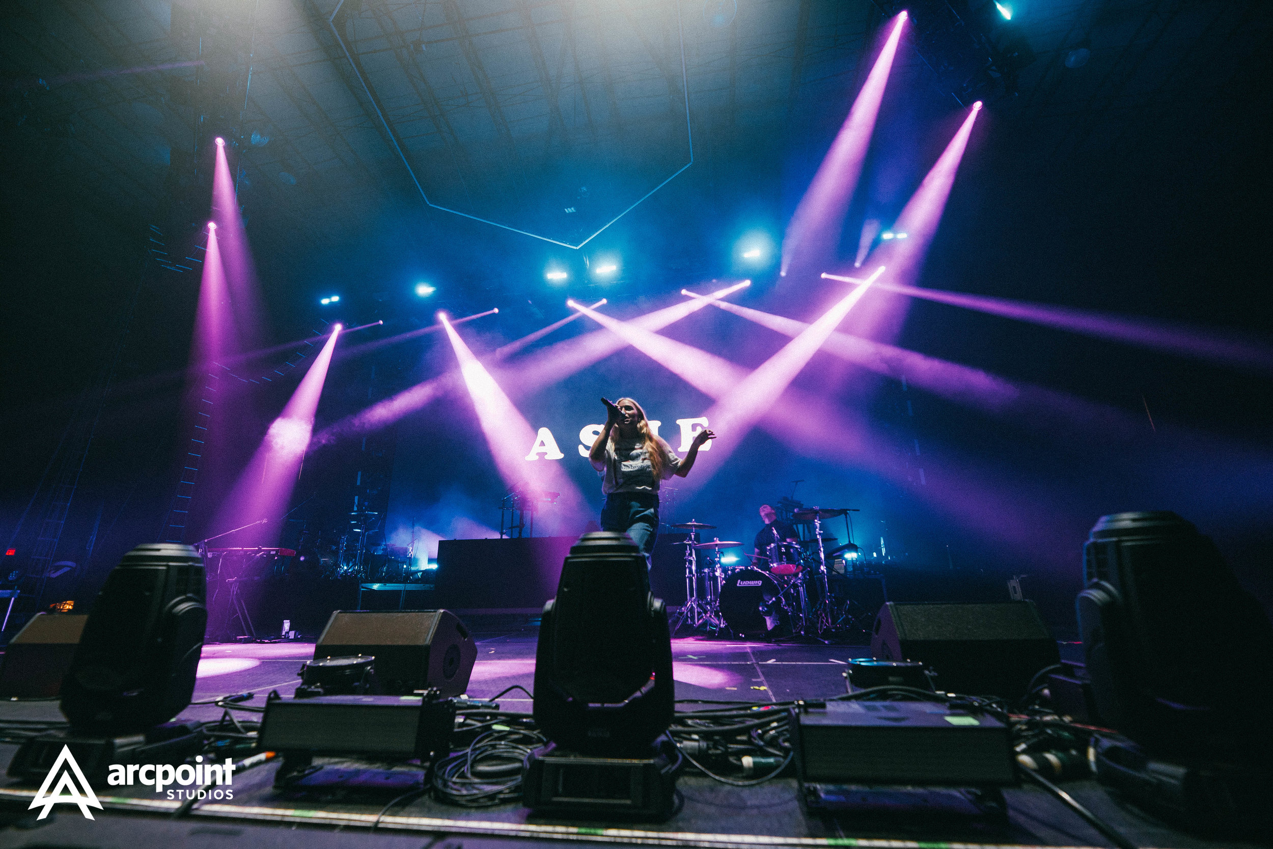 A female singer is performing on stage with purple and blue lighting, backed by live band instruments, in front of a screen displaying the letters 'A S E'.