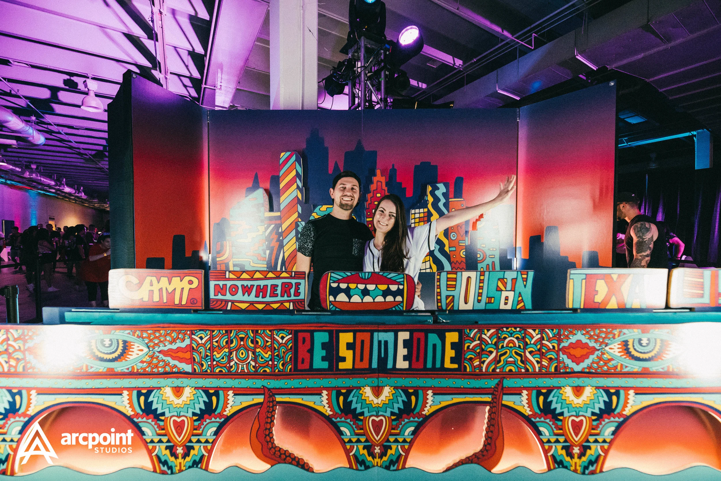 Two people smiling on a colorful stage with artwork and cityscape background at an event. The stage has text reading 'CAMP, NOWHERE, HOUSTON, TEXAS, BE SOMEONE'. There are people in the background and stage lights overhead.