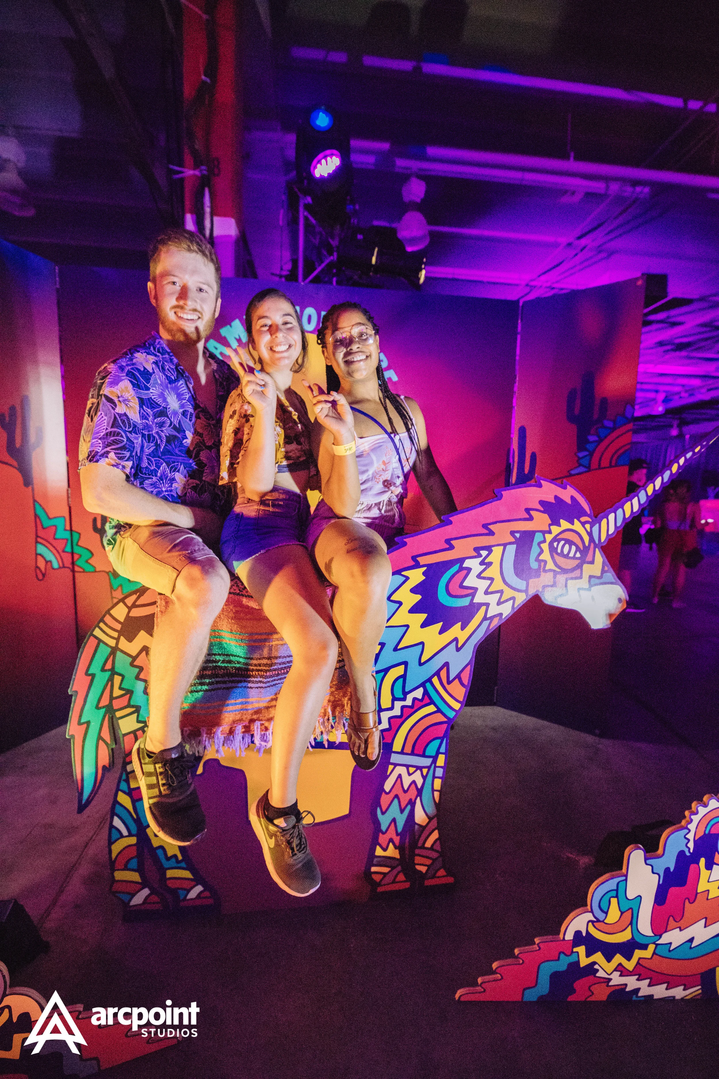 Three friends sitting on a colorful unicorn cutout at a neon-lit event, making peace signs and smiling.