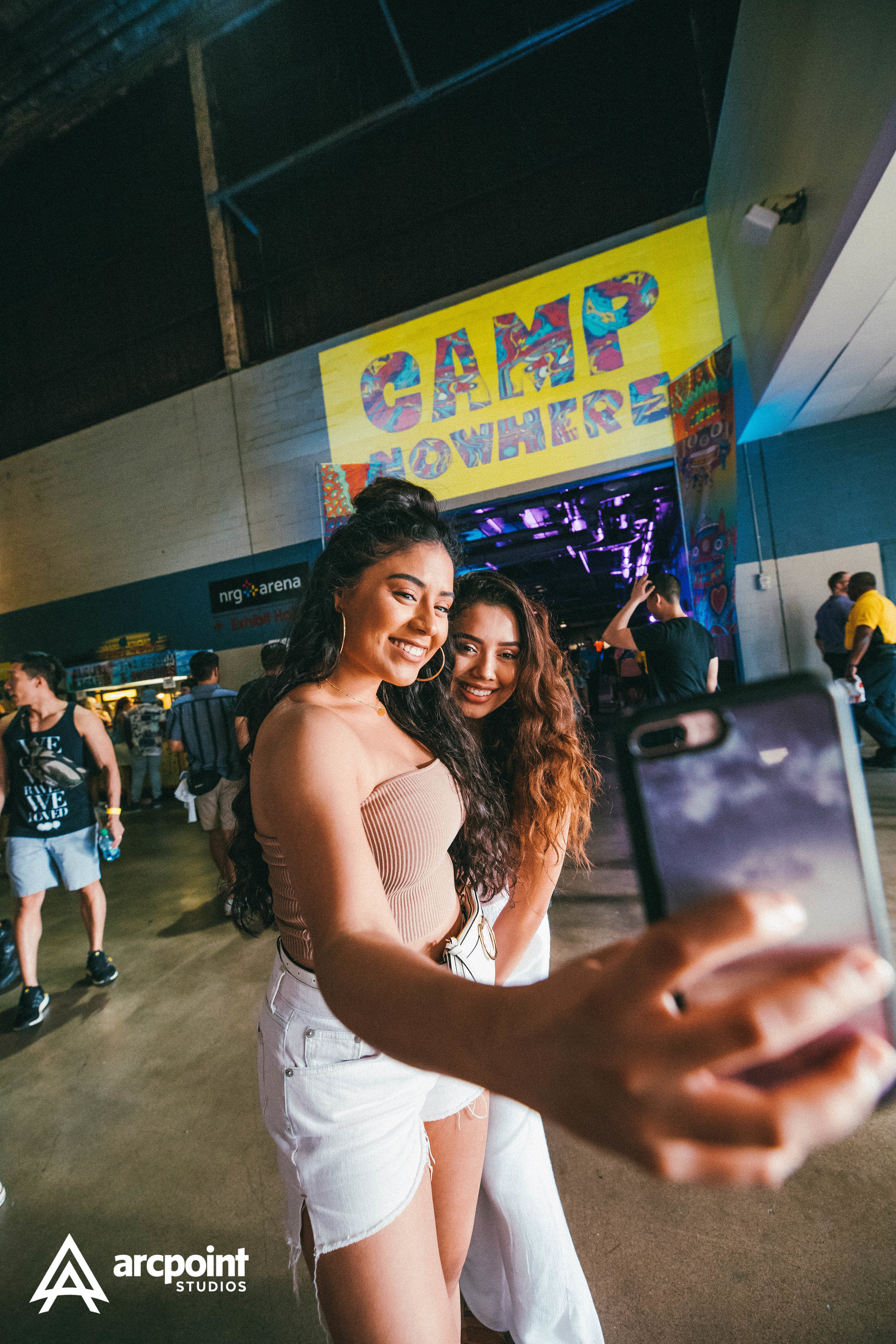 Two young women smiling and taking a selfie at a concert or festival, with a sign that reads "Calif Nowhere" in the background and other people around.