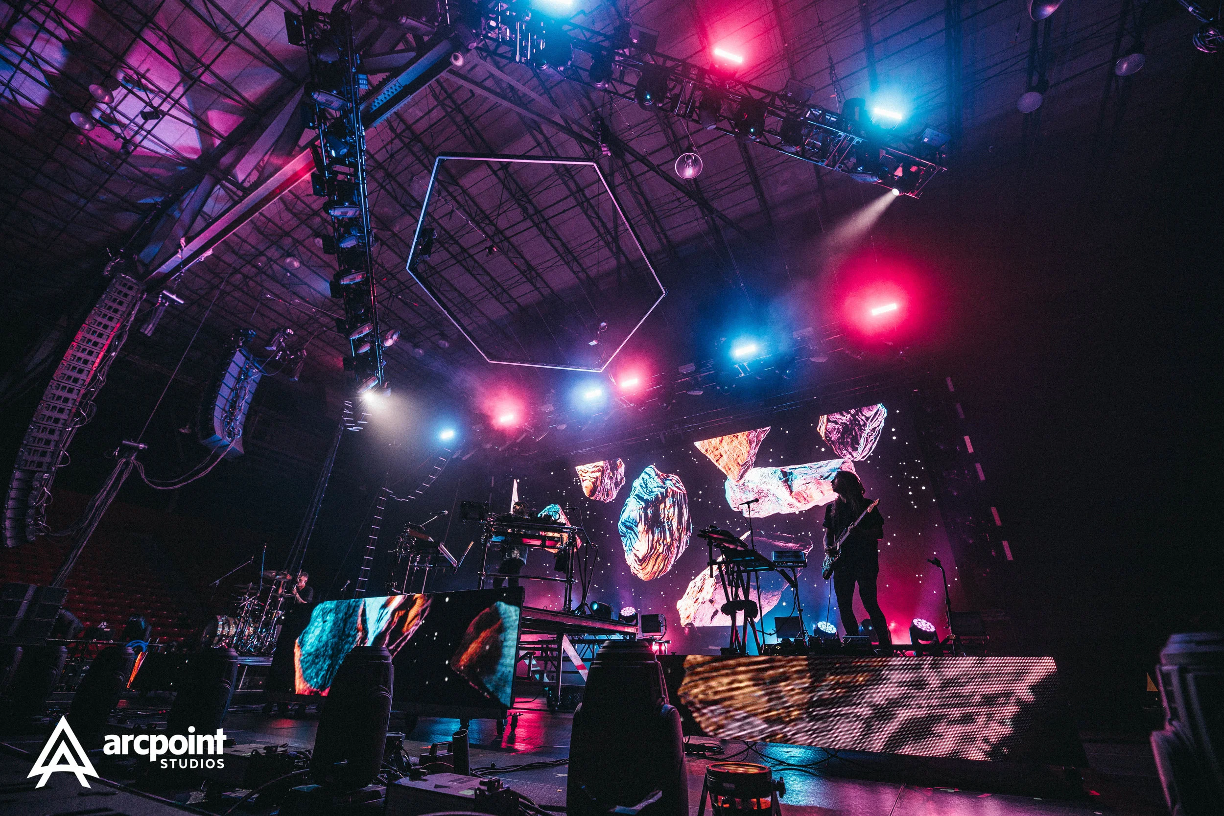 Stage with musicians performing, colorful LED screens showing abstract visuals, vibrant pink and blue lighting, and stage equipment.