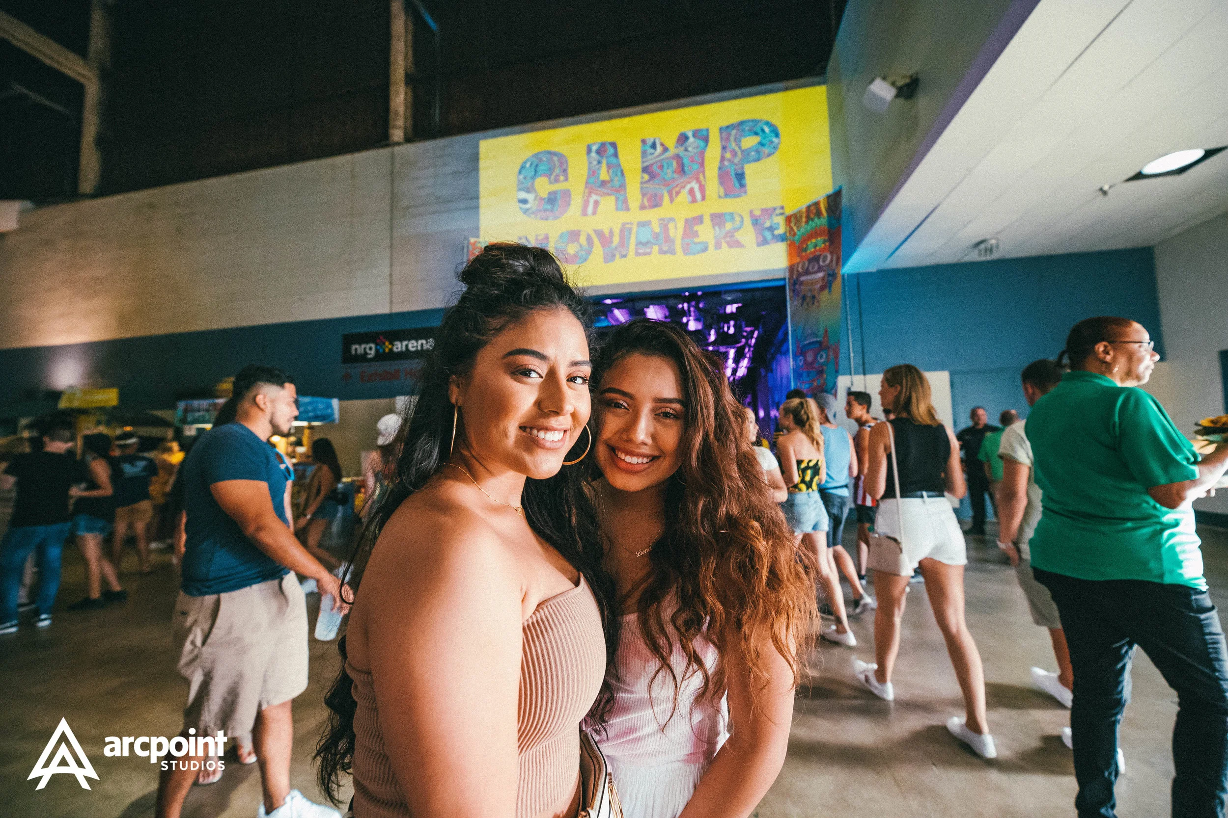 Two young women smiling at the camera inside a lively indoor venue with a crowd of people in the background and a large yellow sign that reads 'CAMP WHERE' above an opening with purple lighting.