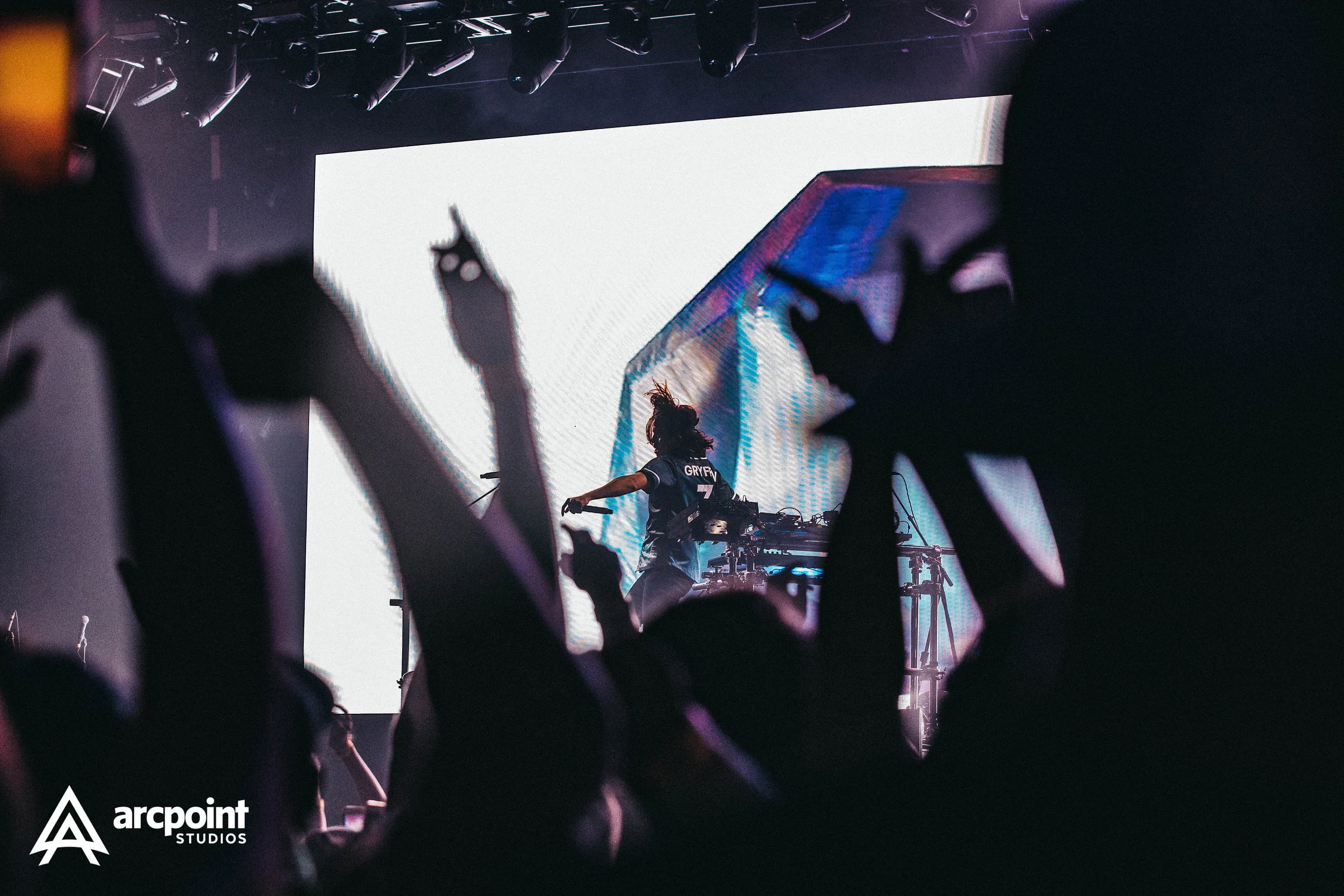 DJ performing on stage at a concert, seen through audience silhouettes with hands in the air, large screen in the background, dim lighting, and Arcpointe Studios logo in the bottom left corner.