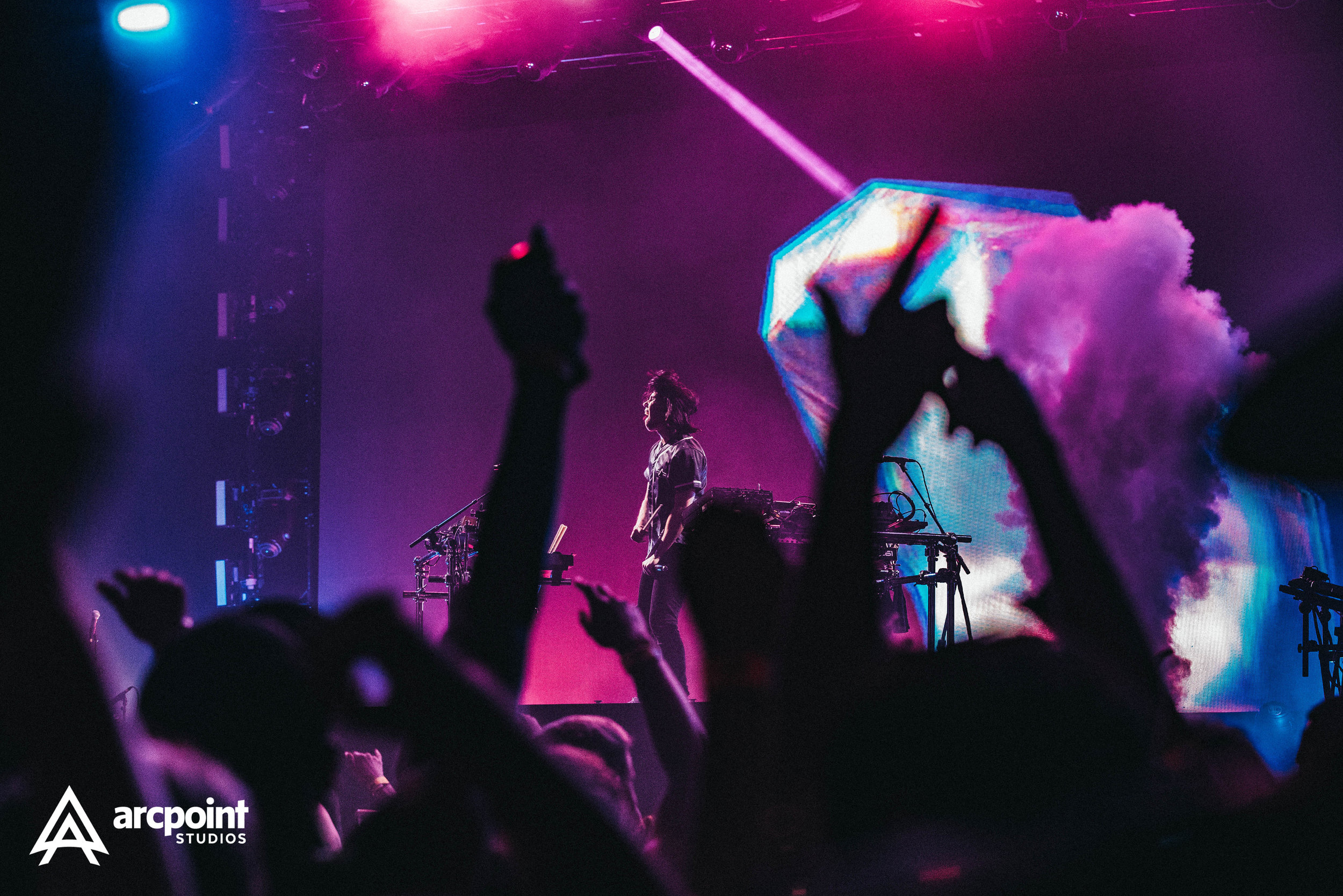 A concert scene featuring a DJ or electronic music performer on stage, surrounded by colorful lights and fog, with audience members raising their hands and taking photos.