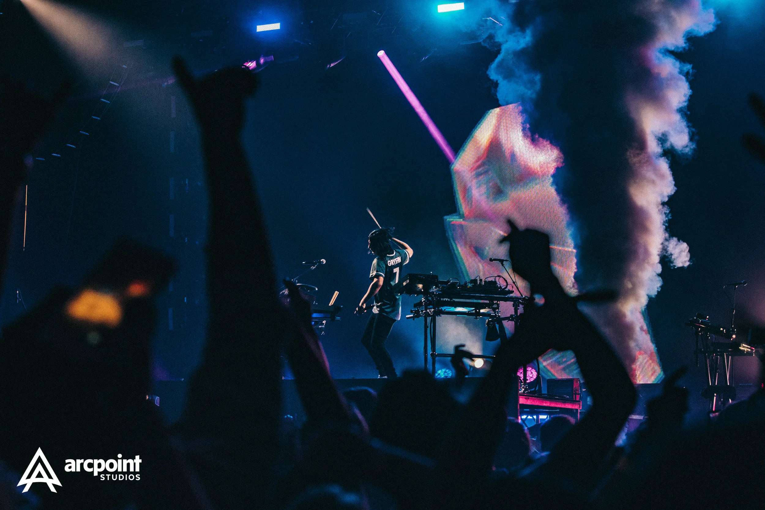 Concert scene with a performer on stage wearing a sports jersey, surrounded by silhouetted audience members raising their hands, colorful lights, smoke, and a large screen displaying visuals.