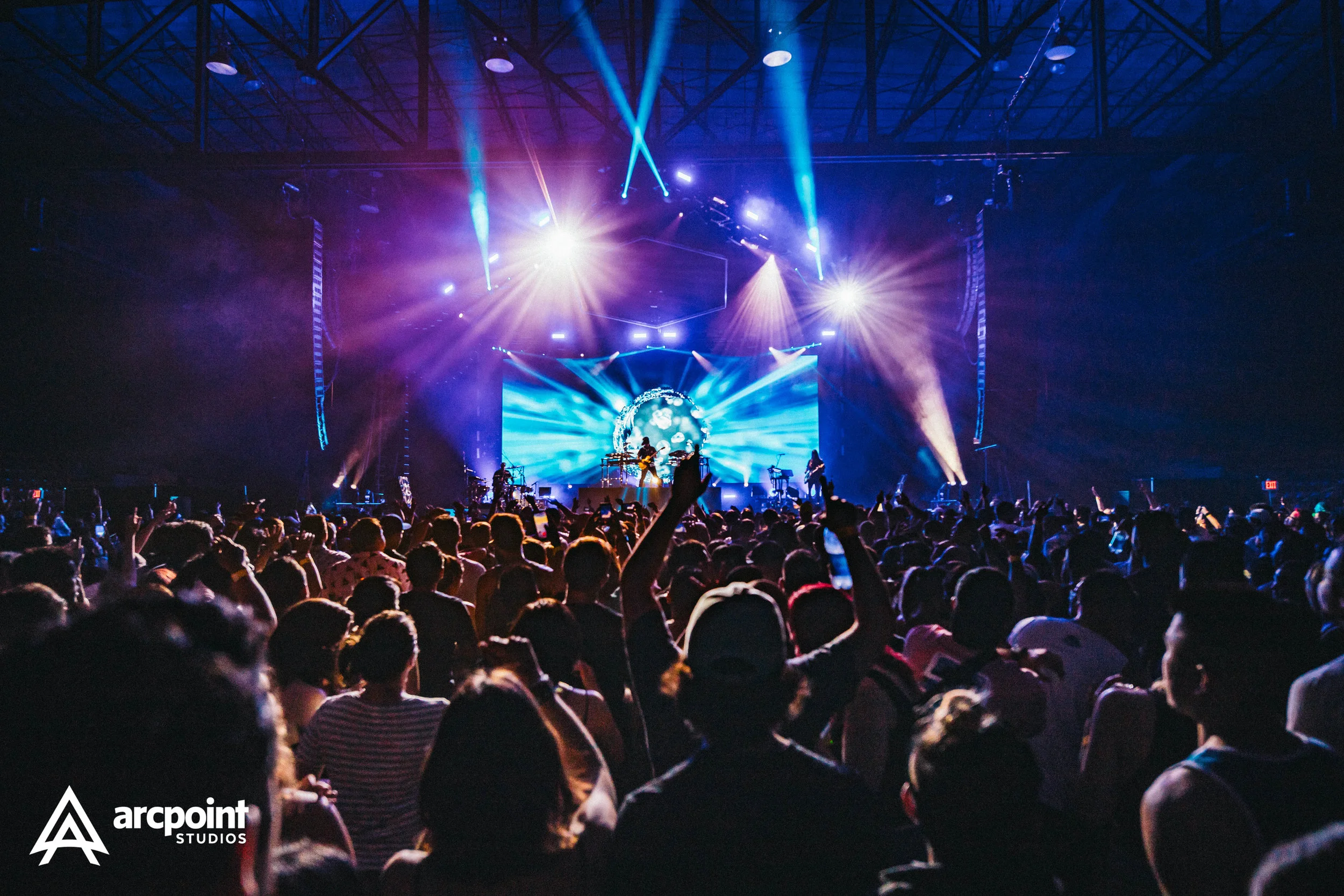 Concert crowd facing stage with bright blue and purple lights, large screen with visual display, and audience members raising hands.
