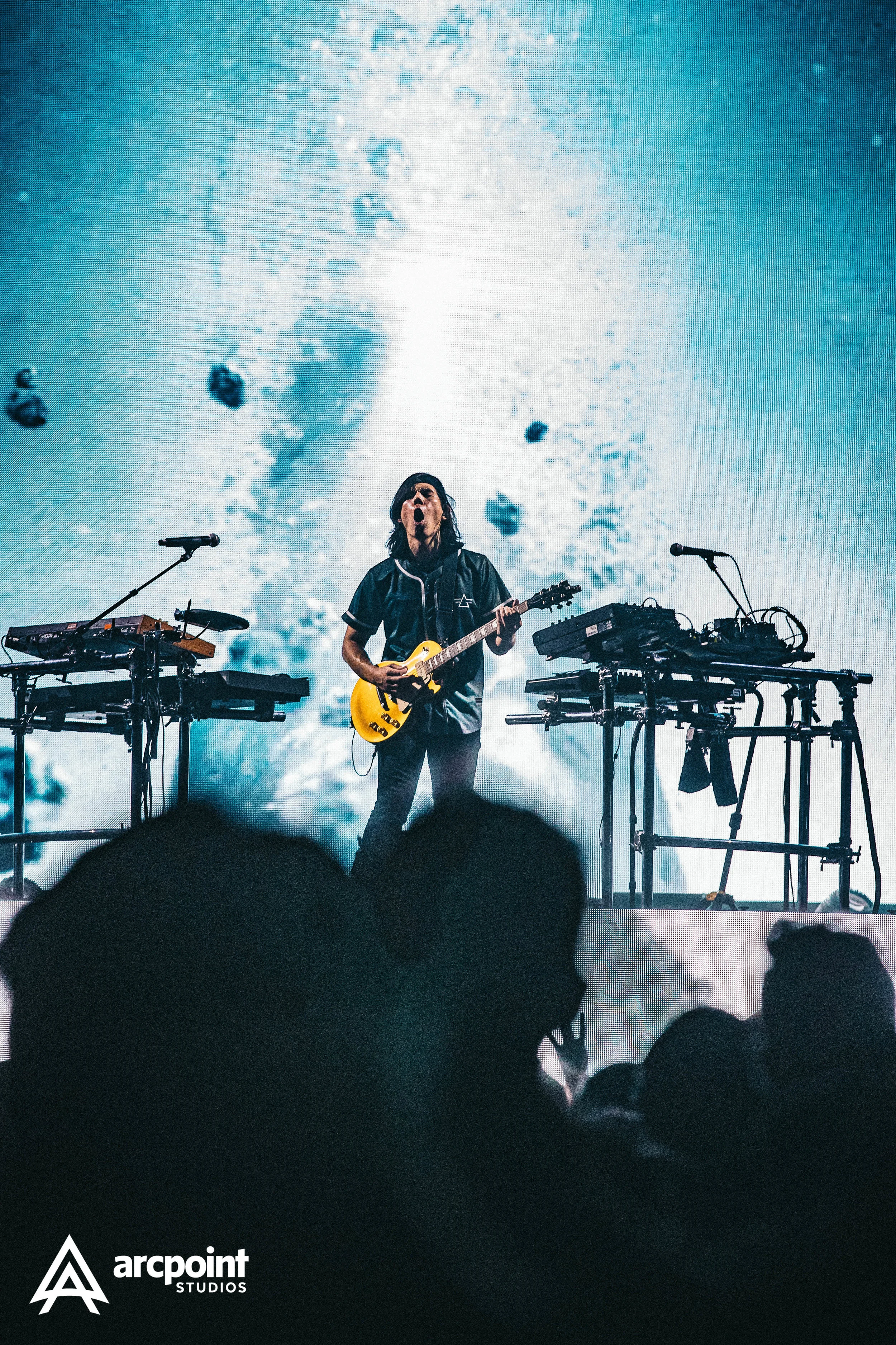 A musician performing on stage with a guitar, surrounded by electronic music equipment, with a large video screen behind displaying abstract blue and white visuals.
