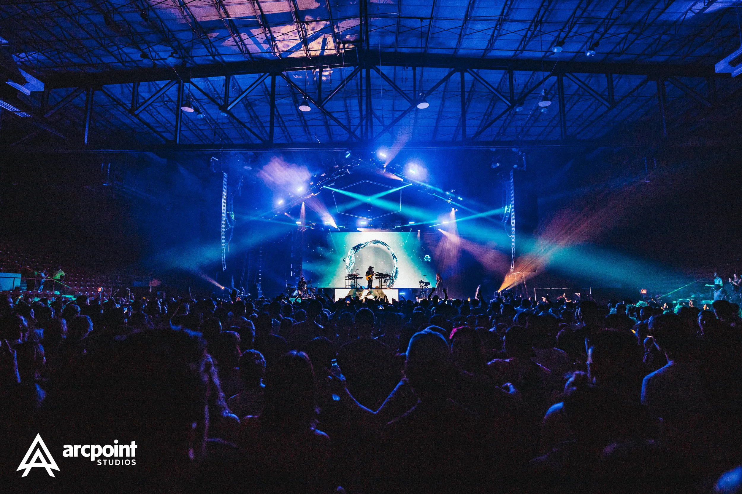 Concert scene with stage, colorful lights, band playing, and audience in a large indoor venue.