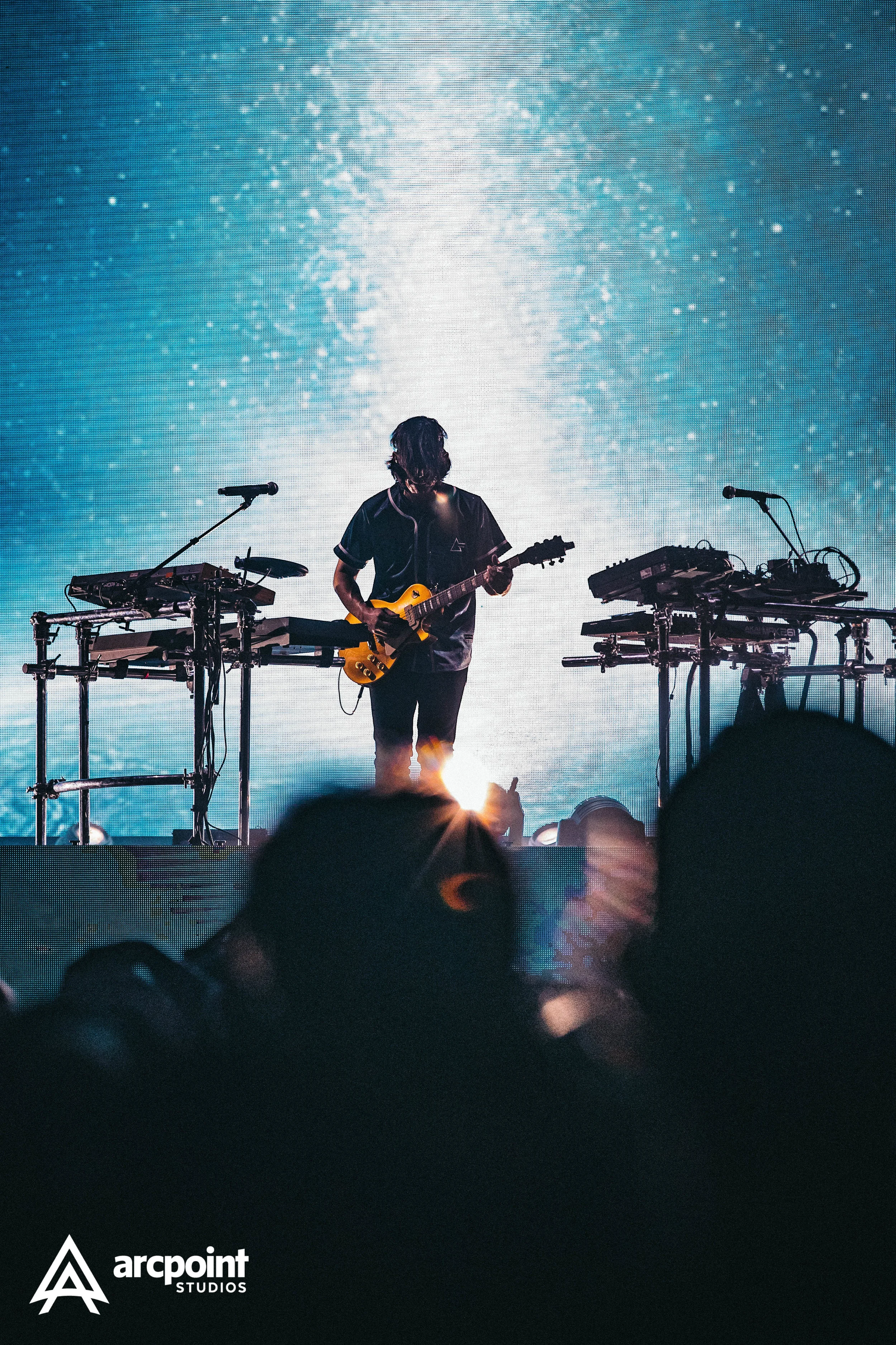 A musician playing an electric guitar on stage with electronic equipment around him, silhouetted against a large digital screen displaying blue and white abstract visuals, with an audience in the foreground.