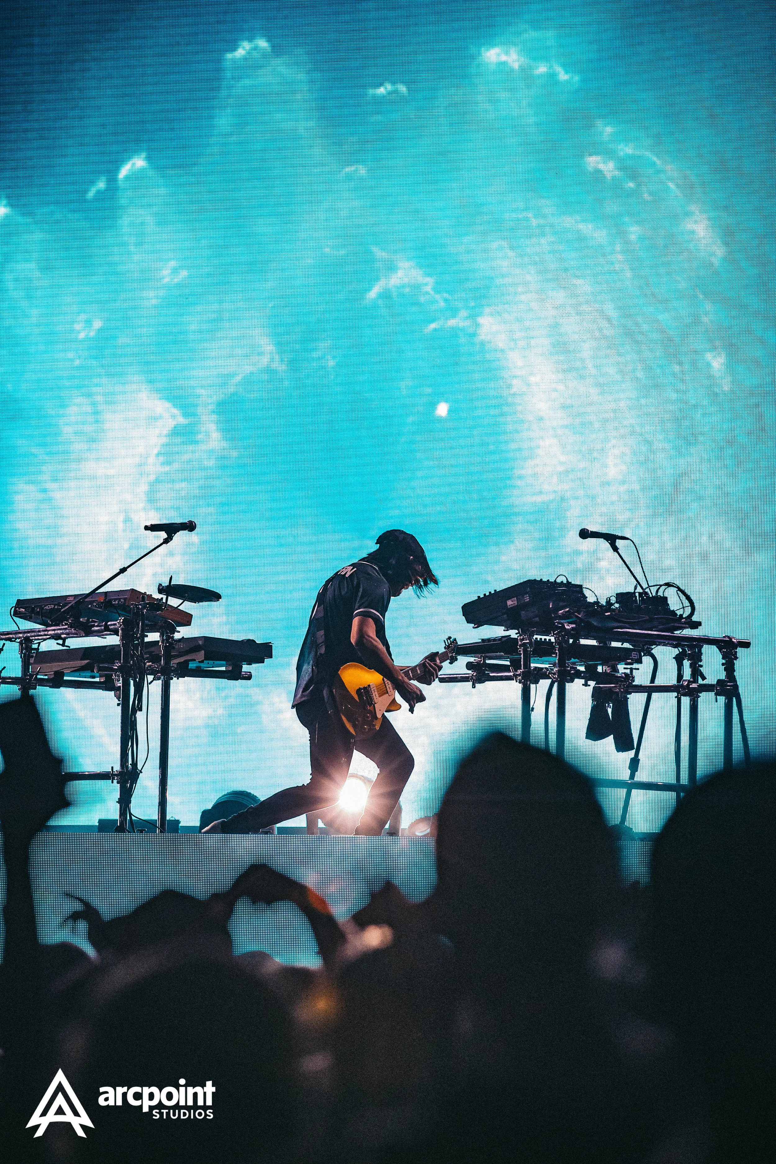 A musician playing guitar on stage with electronic instruments in front of a large illuminated screen at a concert.