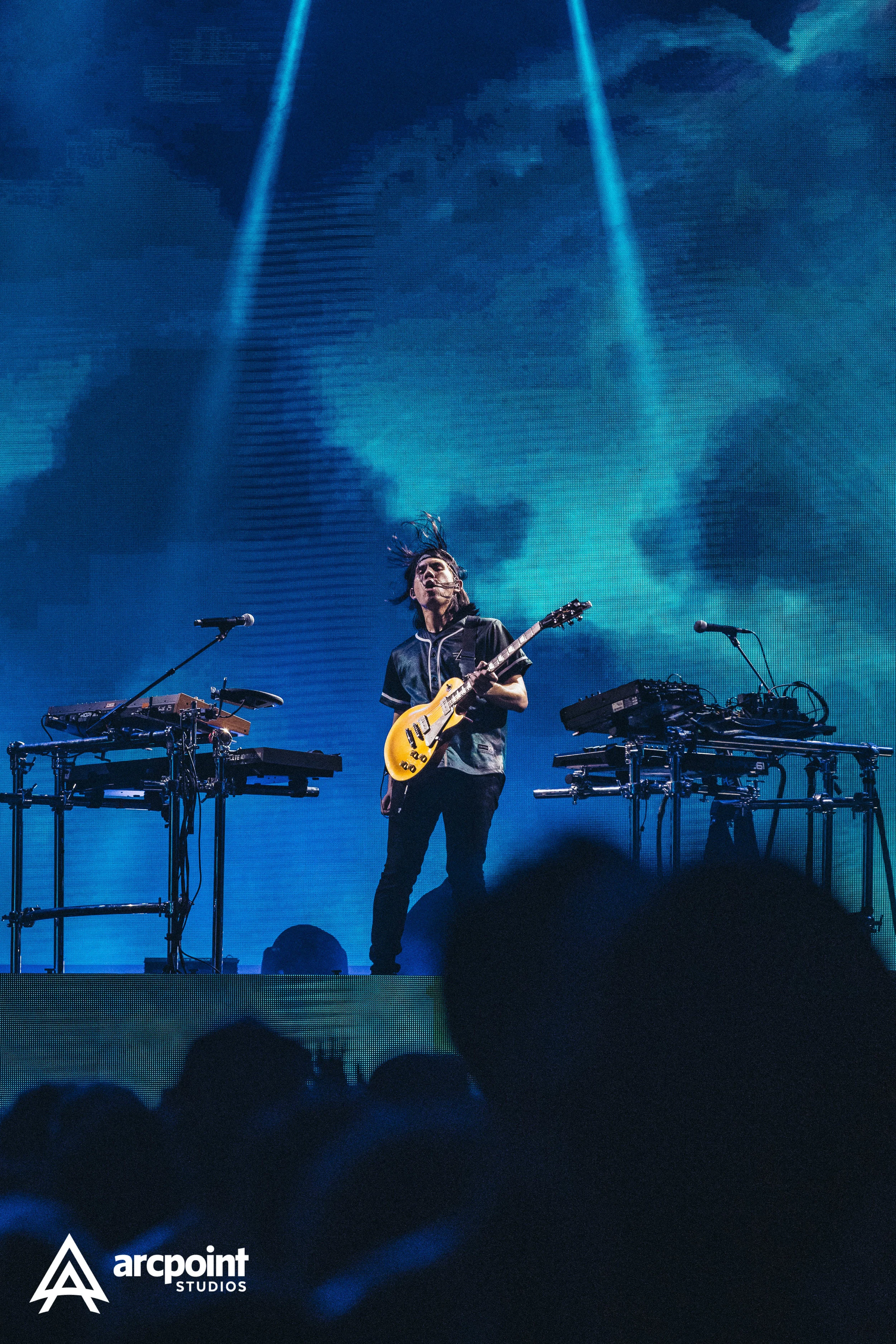 A musician performing on stage with a yellow electric guitar, surrounded by electronic music equipment, against a digital background with blue lighting and cloud patterns.