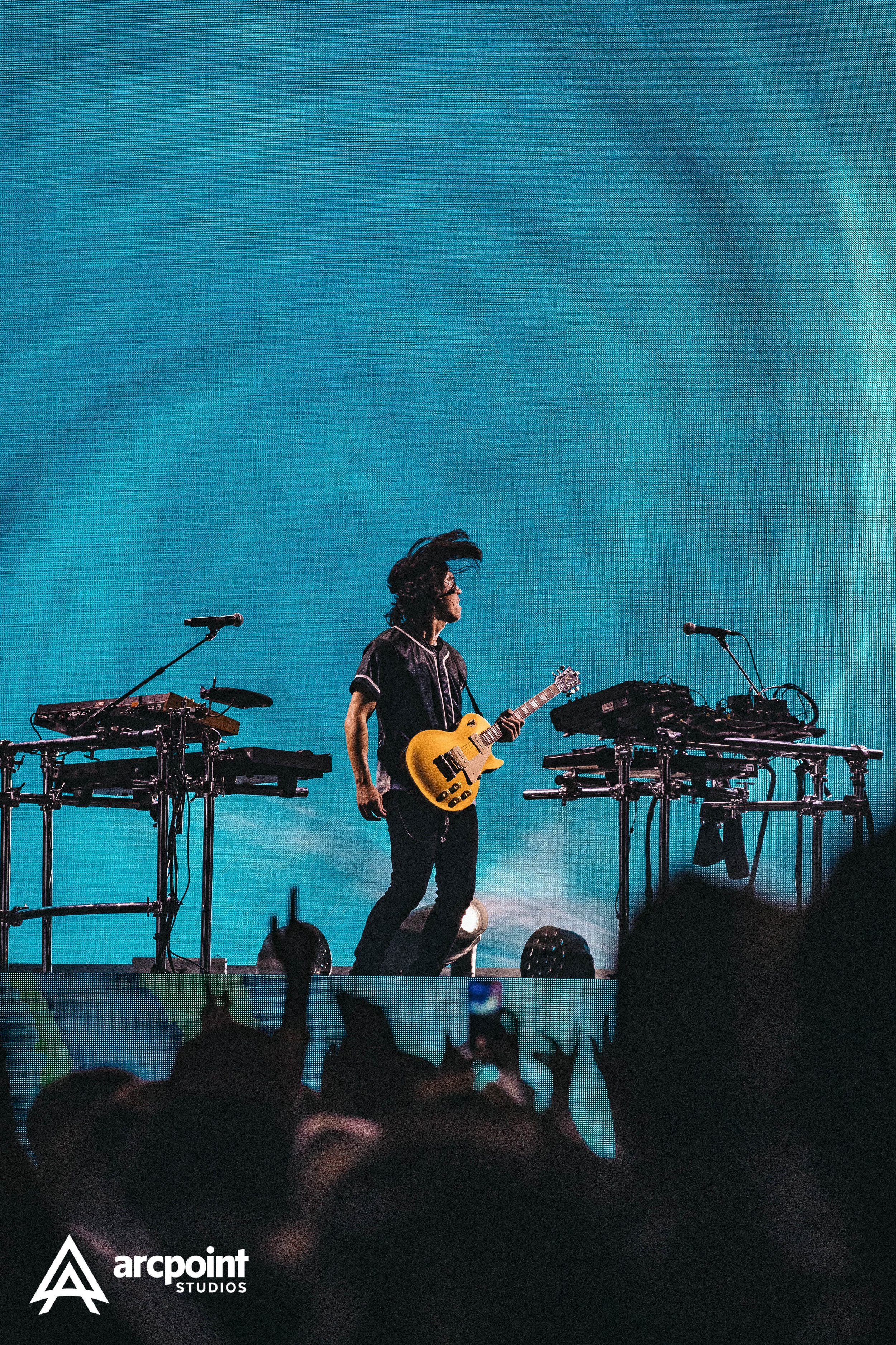 Musician playing yellow electric guitar on stage with electronic equipment, crowd in foreground, large digital screen in background.