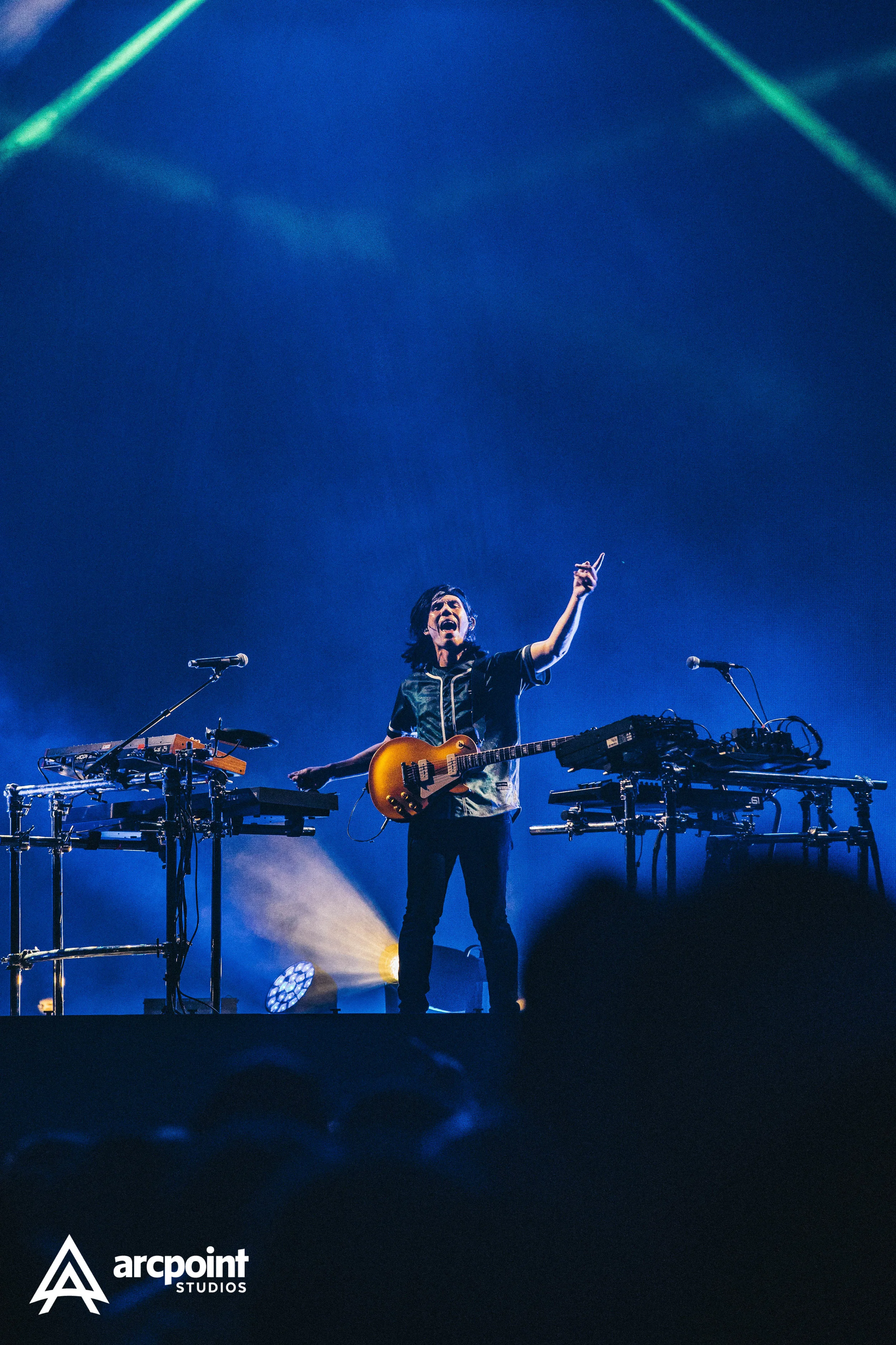 A musician playing an electric guitar on stage during a concert, with electronic equipment and microphones on stands around him, illuminated by blue and yellow stage lights.