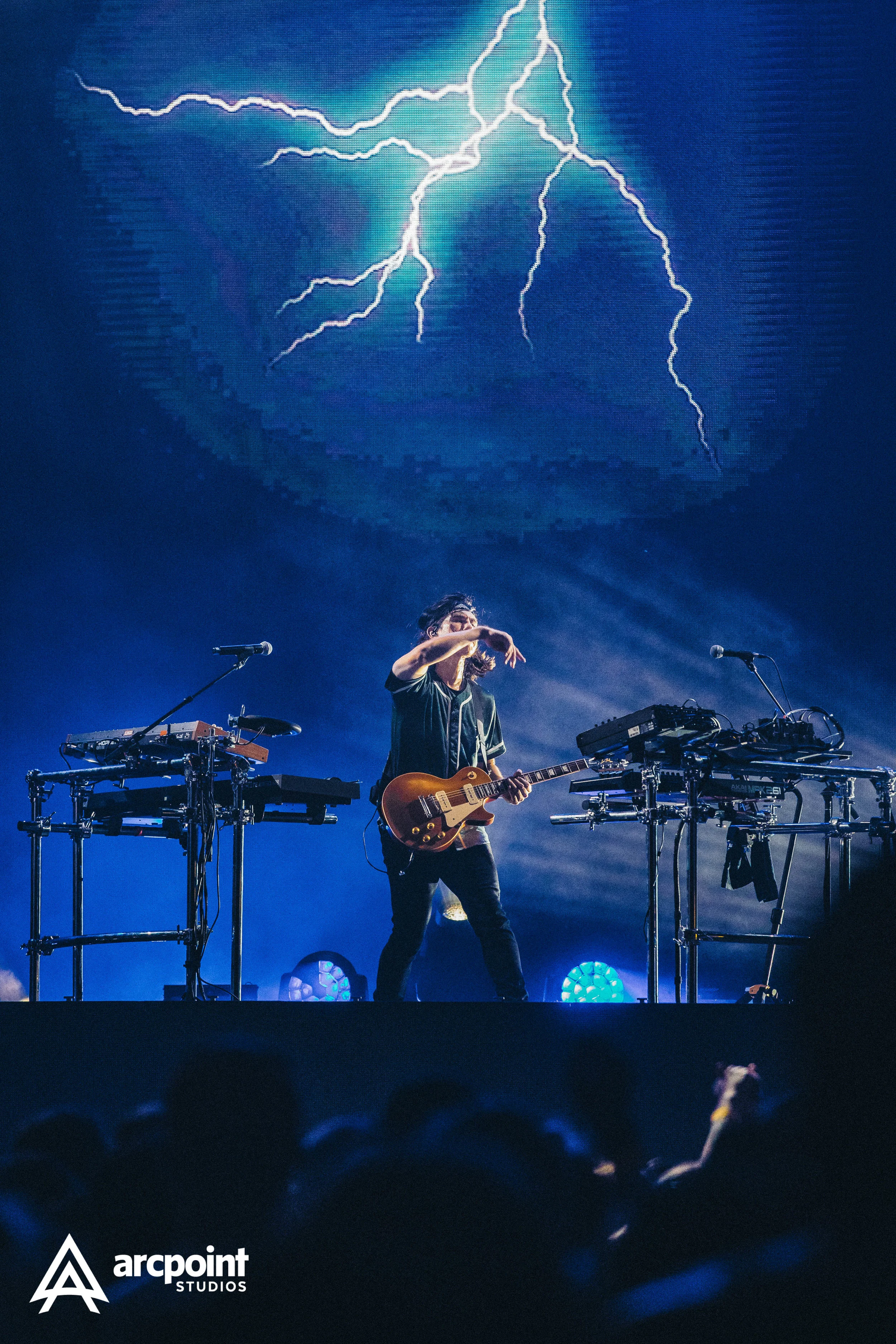 A musician with long hair playing an electric guitar on stage with electronic music equipment nearby. Behind him, a large screen displays a lightning storm image.