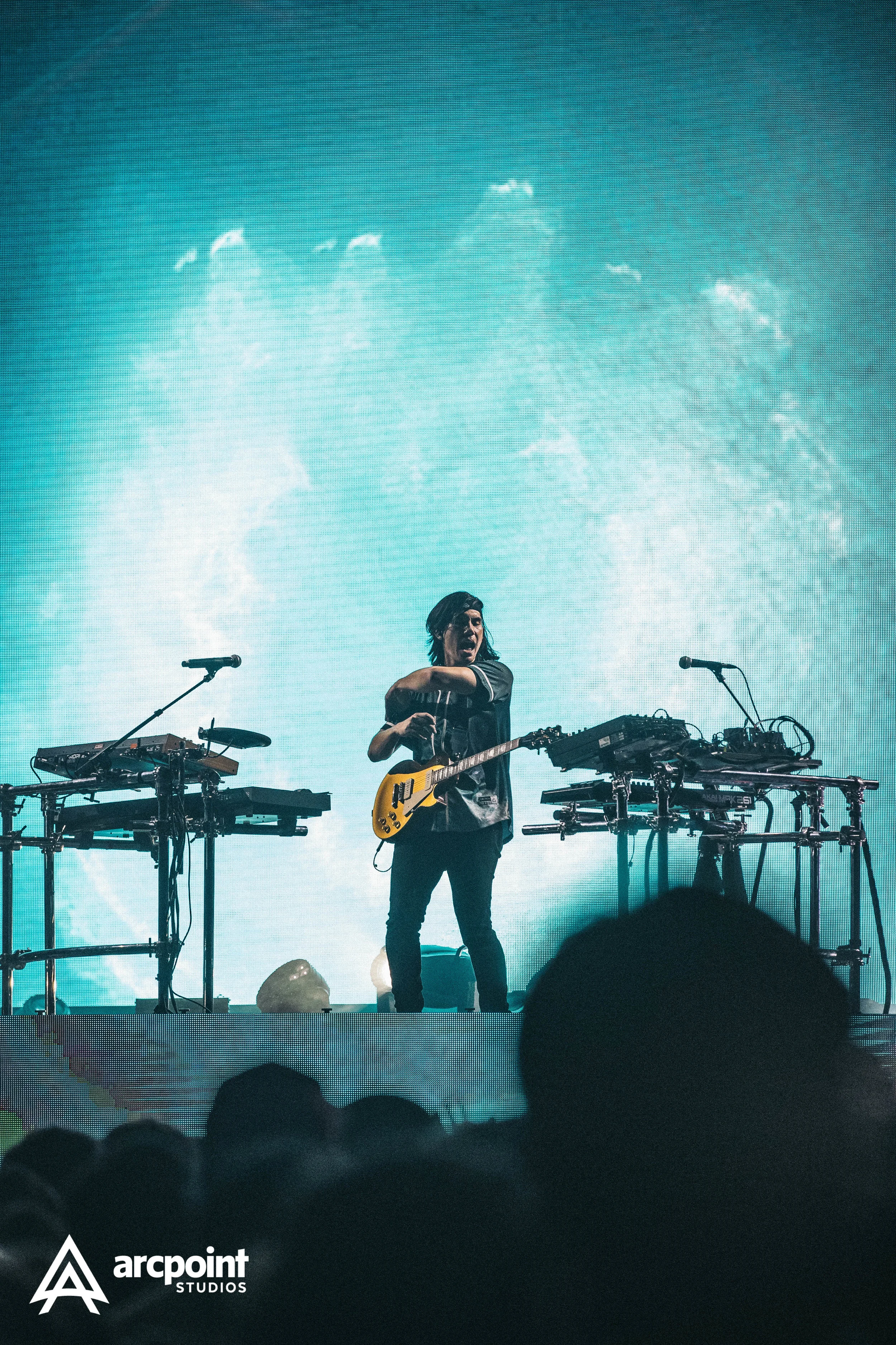 A musician with long hair playing a guitar on stage with electronic music equipment, a large digital screen displaying an ocean wave behind him, and a crowd in the foreground.