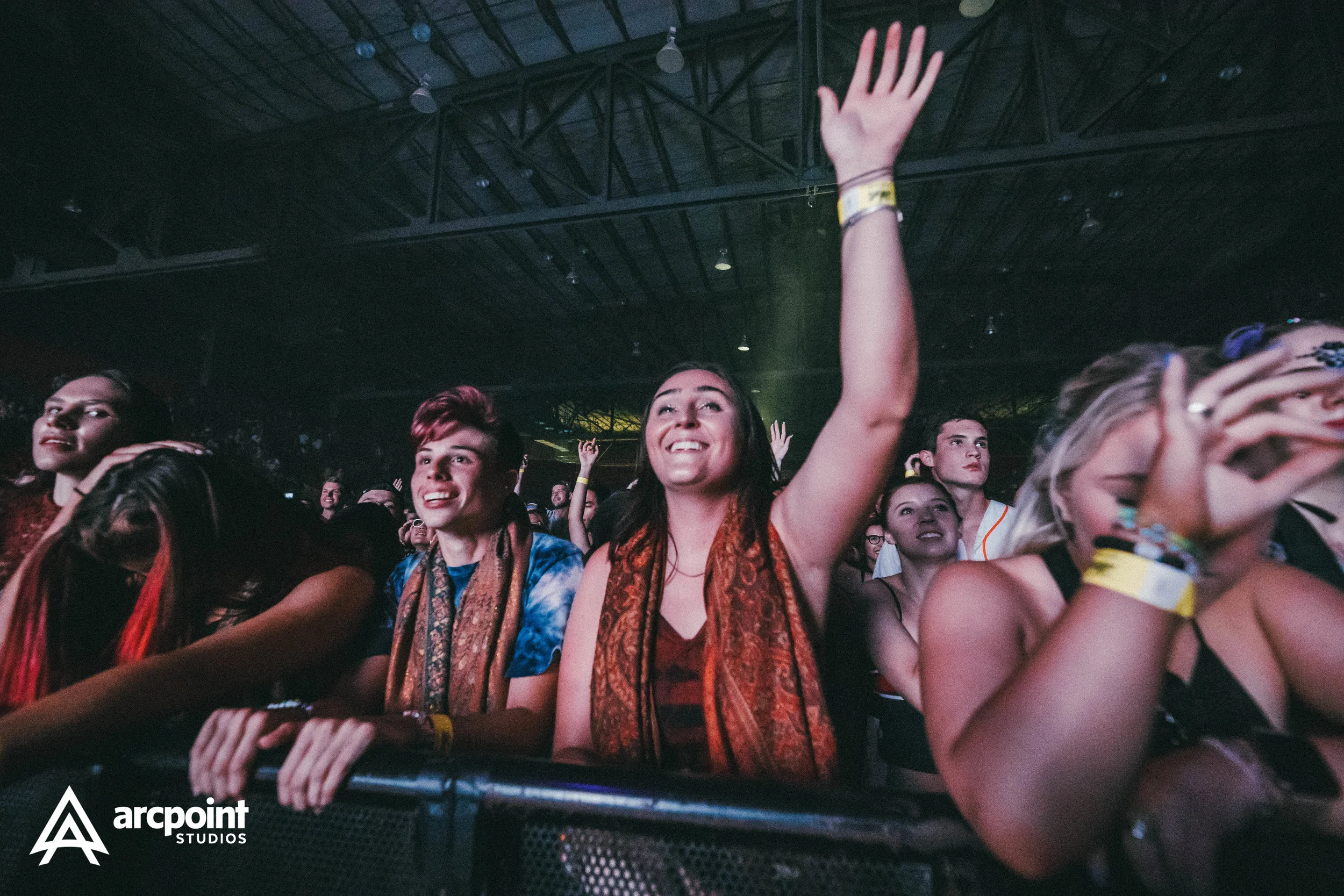 Group of diverse people enjoying a concert, with woman in center raising her hand and smiling.
