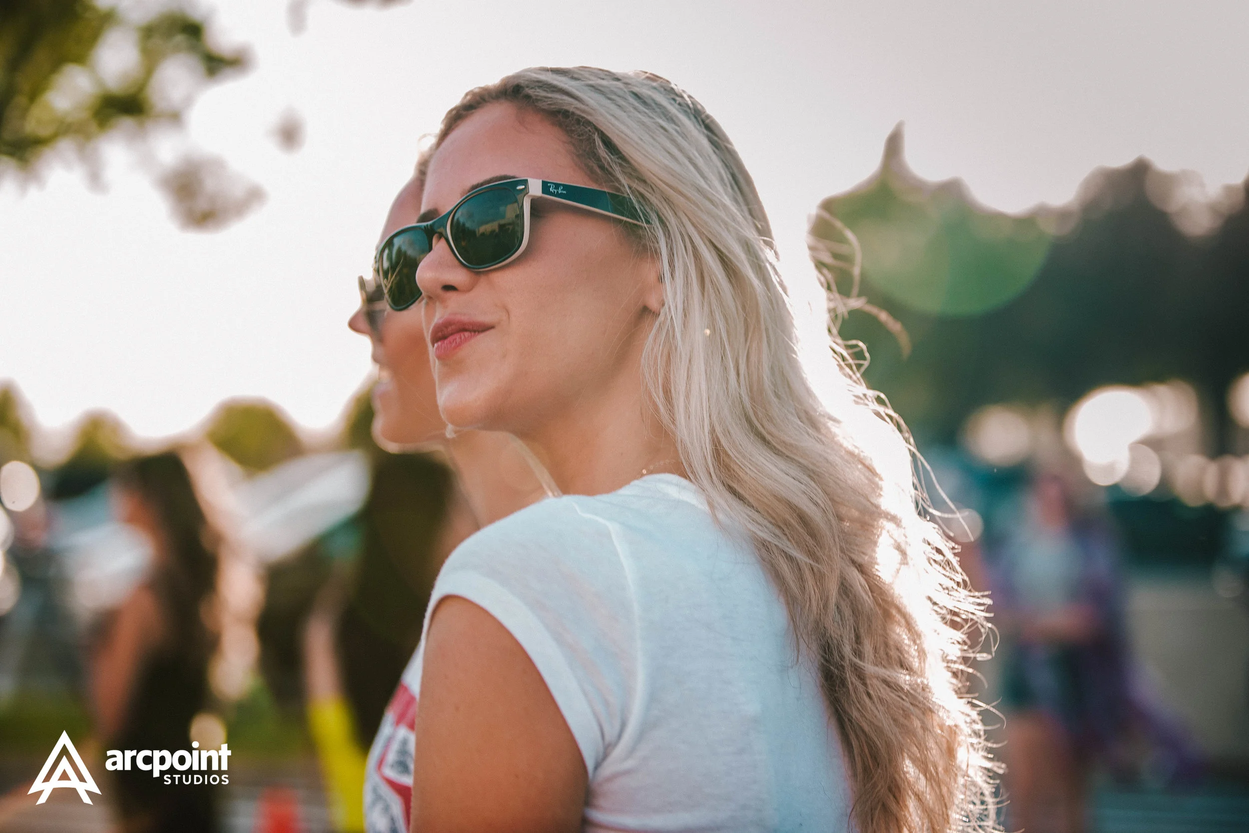 A woman with long blonde hair wearing sunglasses and a white t-shirt, standing outdoors with blurred people in the background.