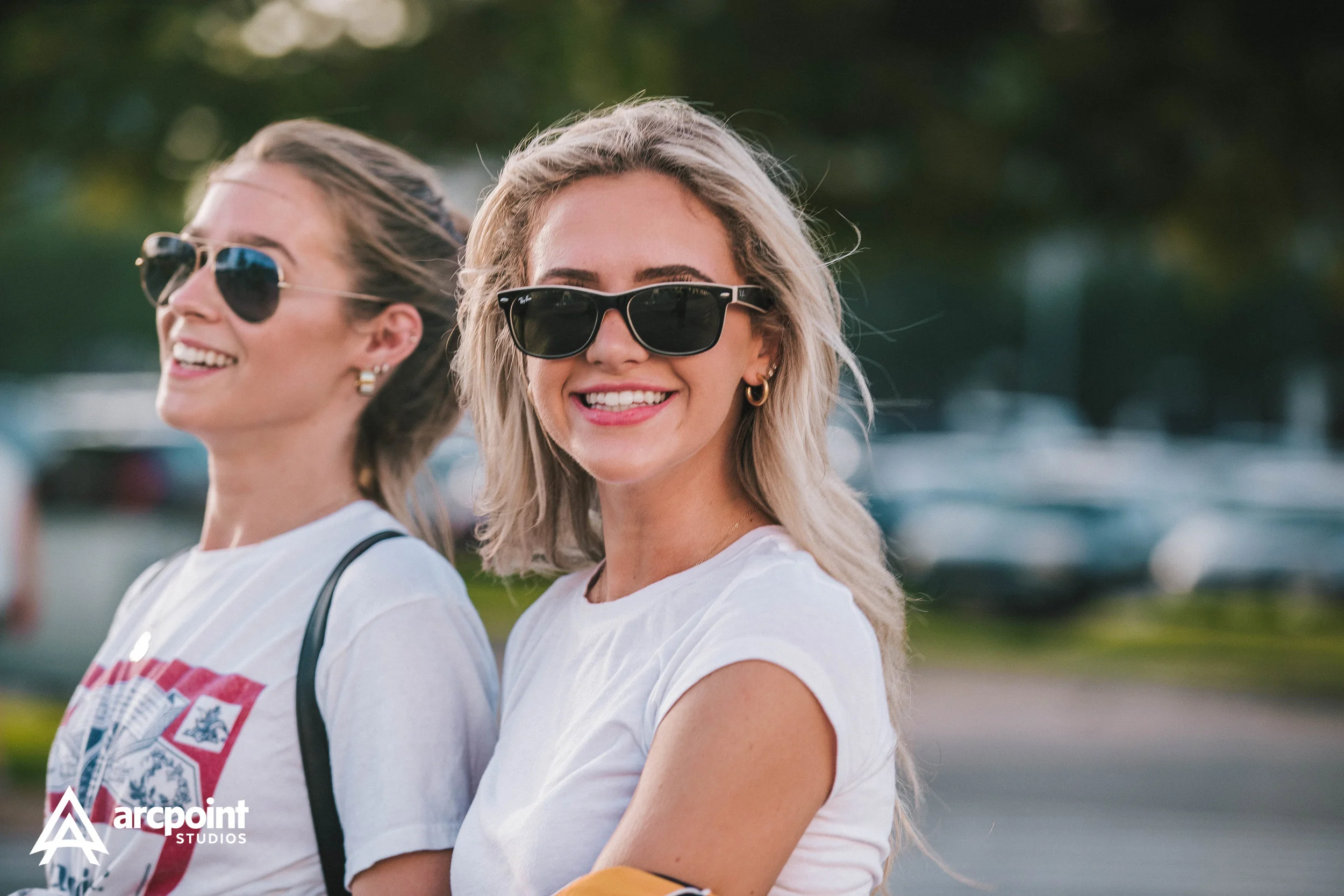Two women wearing sunglasses smiling outdoors, with a parking lot in the background.