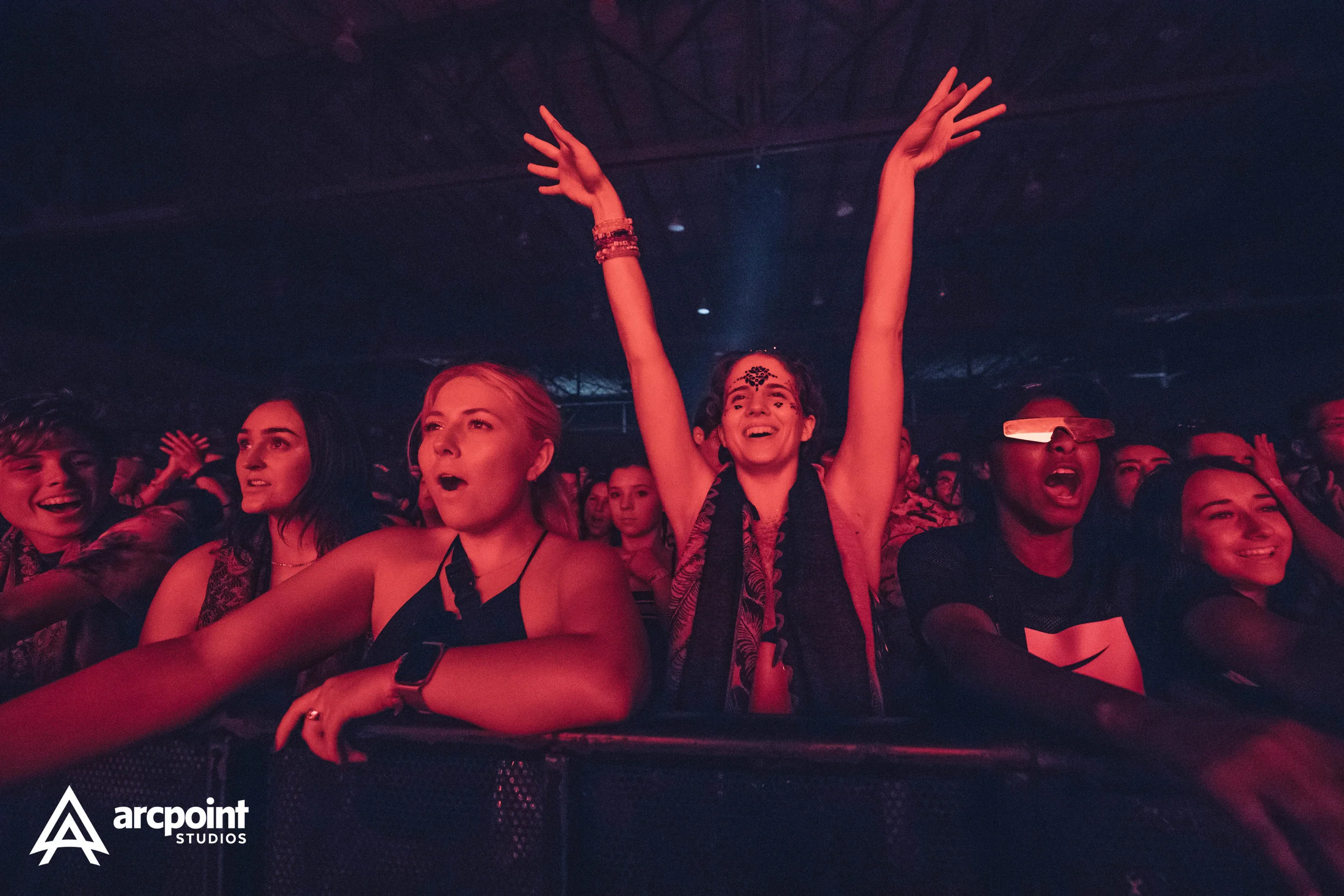 Crowd of young people at a concert, some singing and dancing, one woman in the center raising her arms in excitement, wearing a black scarf and face makeup, with red lighting and a dark background.