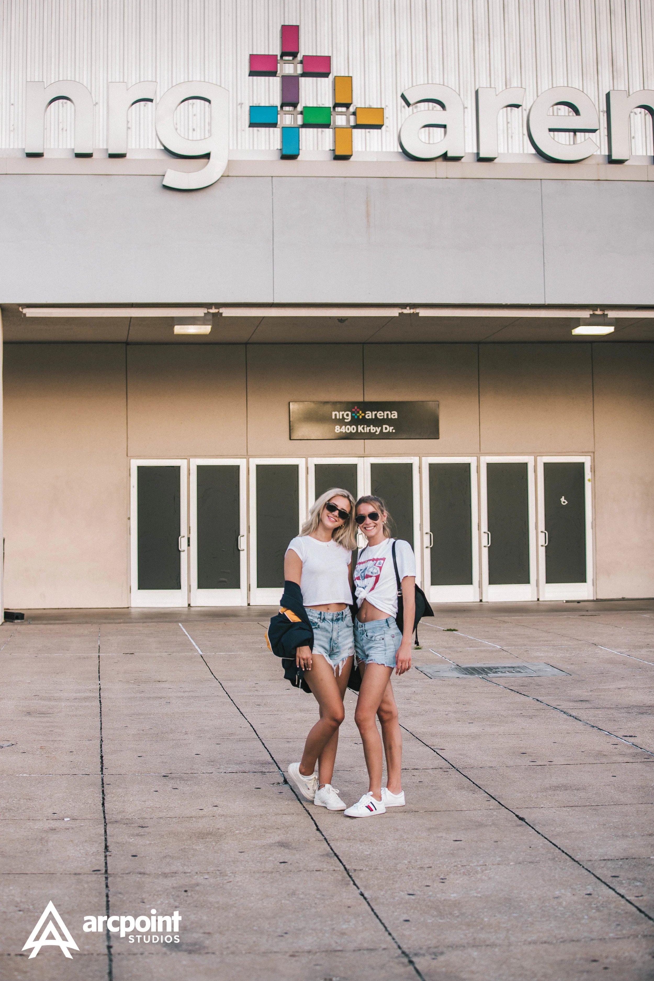 Two young women with sunglasses, wearing casual summer clothes, standing in front of the NRG Arena entrance, smiling at the camera.